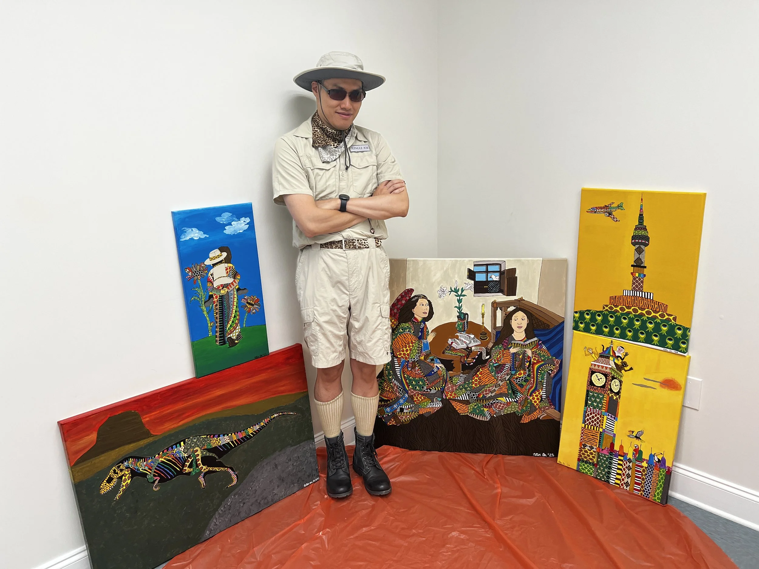 A person wearing a safari hat, sunglasses, and safari clothing standing with arms crossed, surrounded by colorful paintings of African animals and scenes, on a red floor covering against a plain white wall.