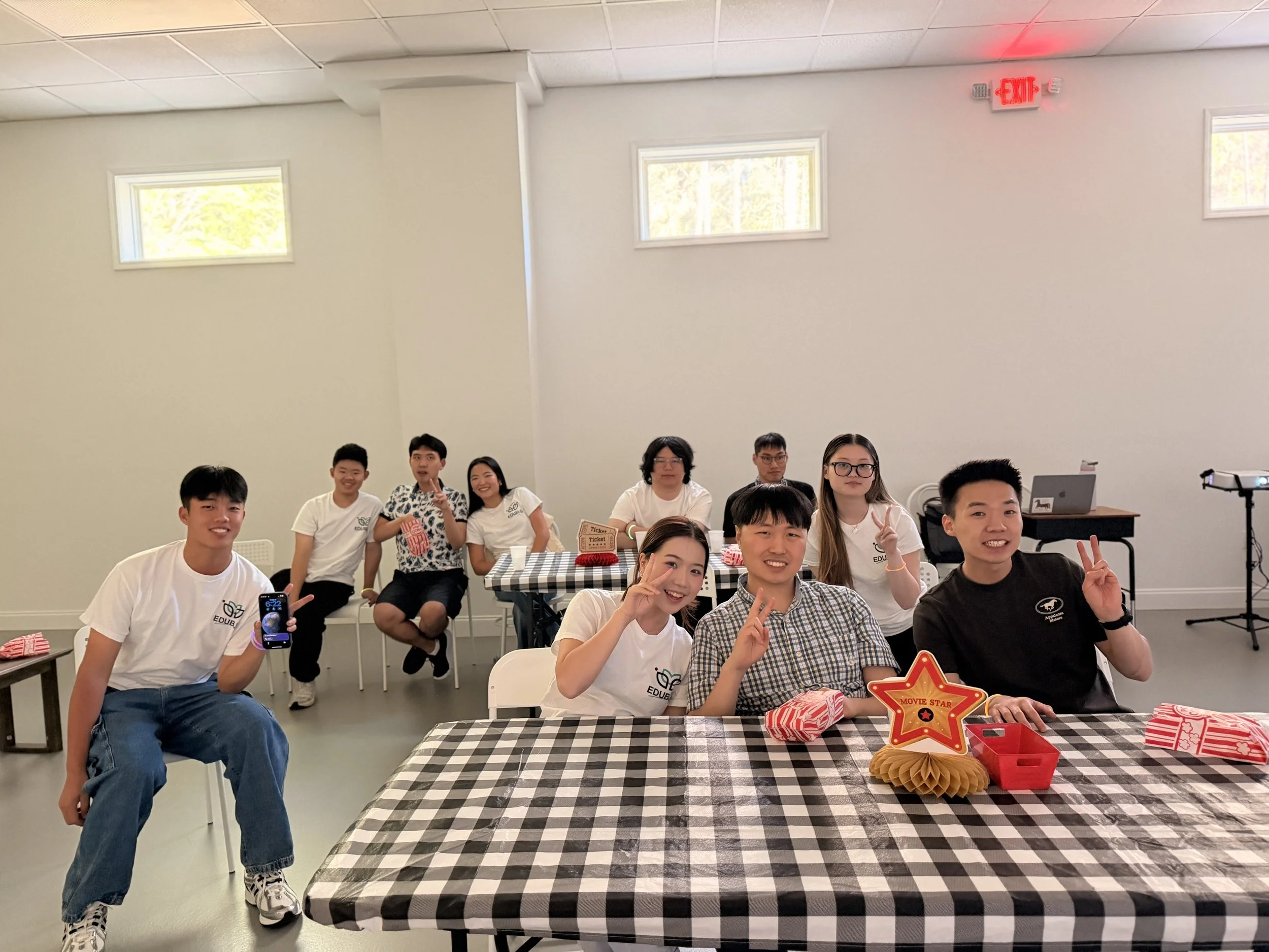 A group of young people sitting and standing around a table with checkered tablecloths during a celebration. Some are making peace signs and smiling at the camera, with party decorations and gift-wrapped boxes on the table. There is a bright, spaciou