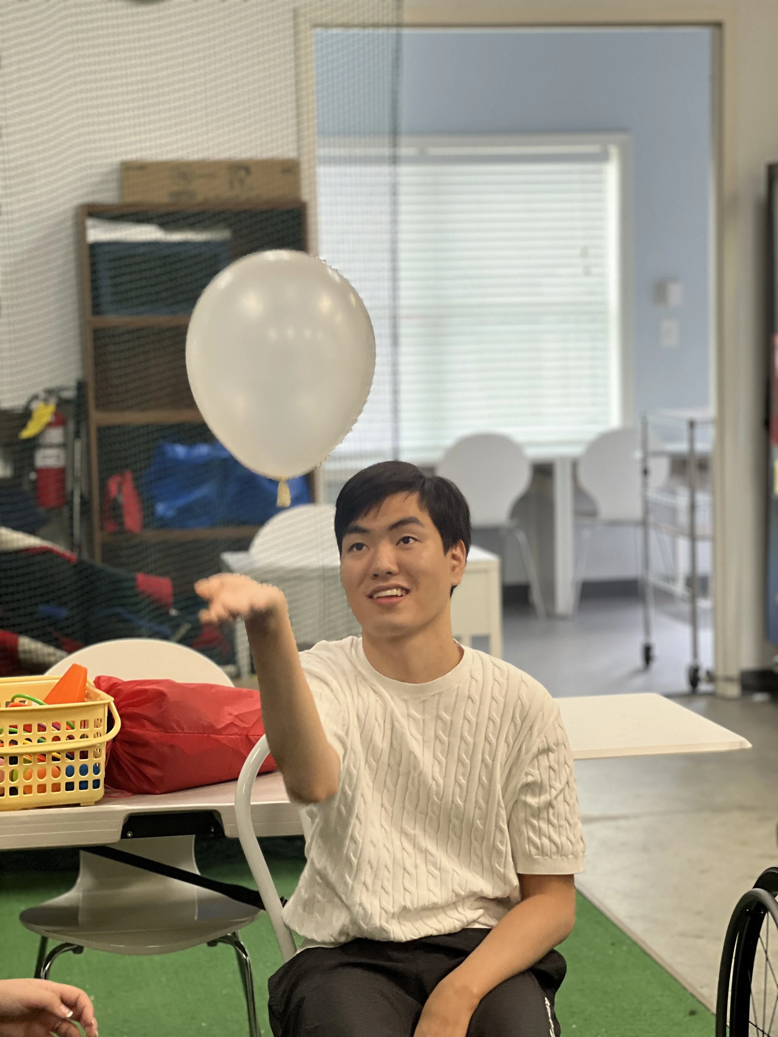 A young man wearing a cream-colored sweater sitting indoors with a white balloon floating above him. There are chairs, a basket with colored toys, a red bag, and other furniture in the background.
