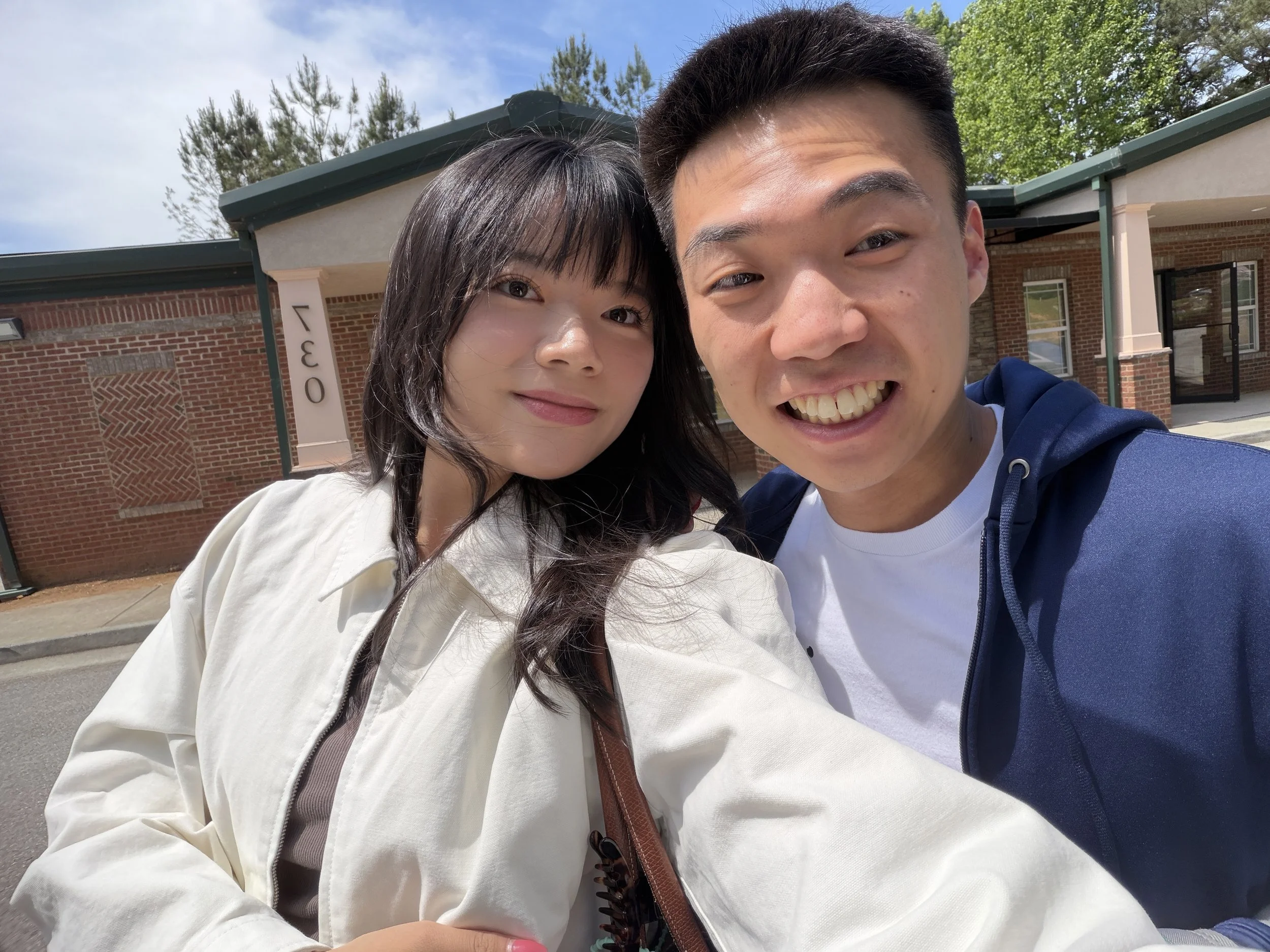 Two young people, a woman with dark hair and a man with short dark hair, taking a selfie outside in front of a brick building and green trees on a sunny day.