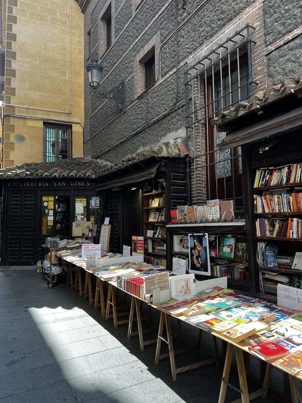 Outdoor street bookstore with tables of books in front of a bookstore named Libreria San Gines, with a building and window with security bars in the background.