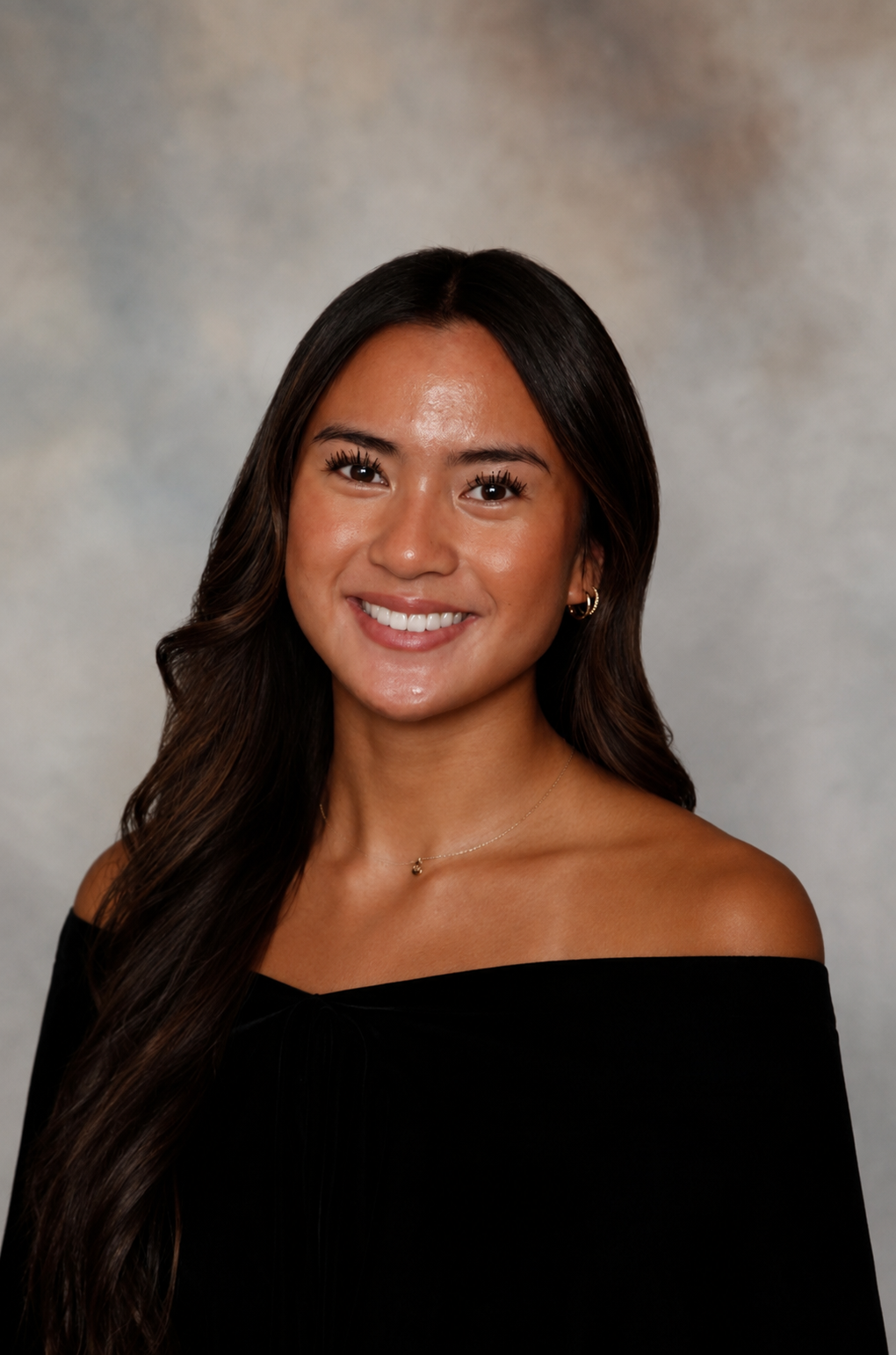 A young woman with long dark wavy hair, smiling, wearing a black off-the-shoulder top, gold earrings, and a delicate necklace, against a gray textured background.