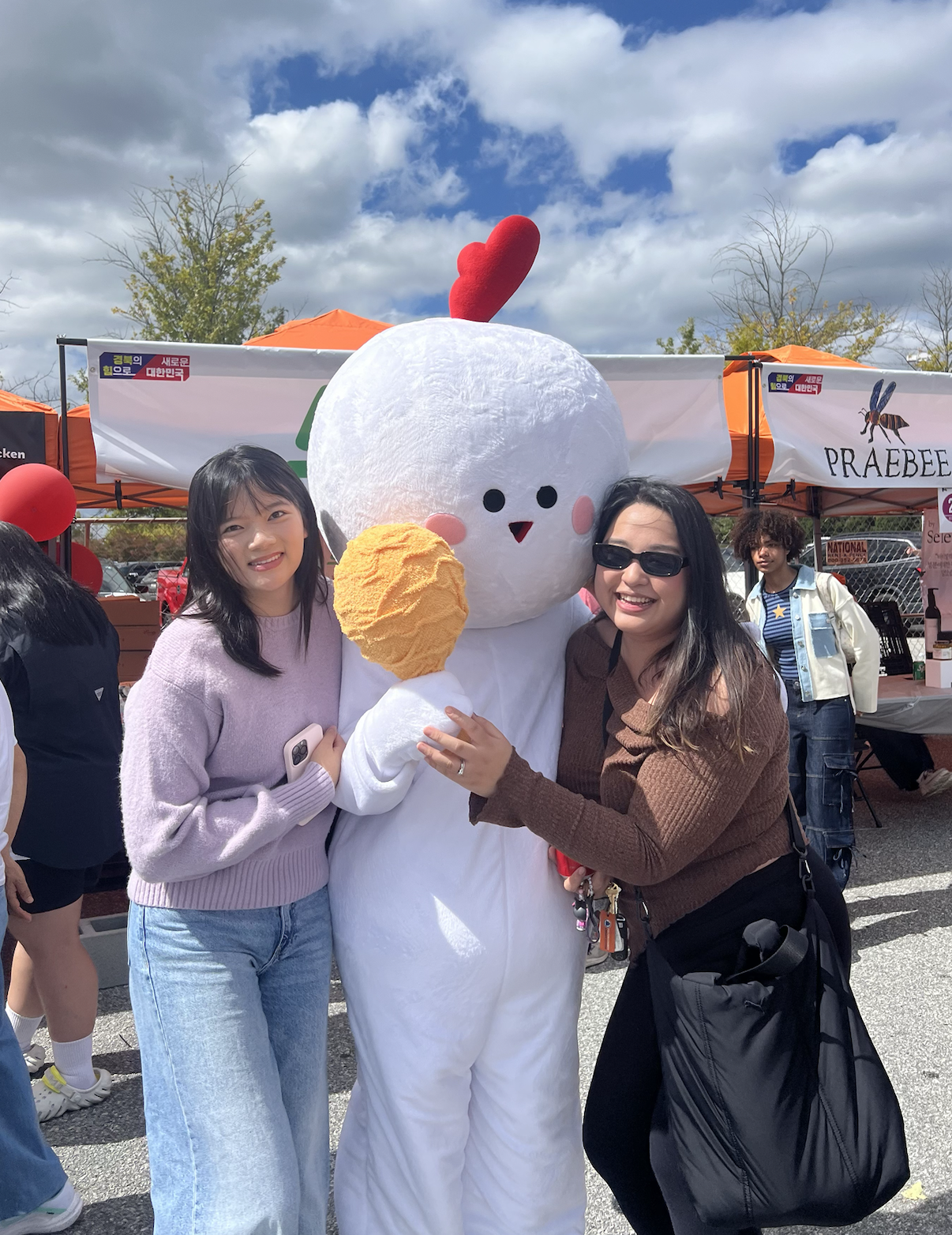 Two women smiling and hugging a person in a large snowman costume with a red heart on its head at an outdoor event.