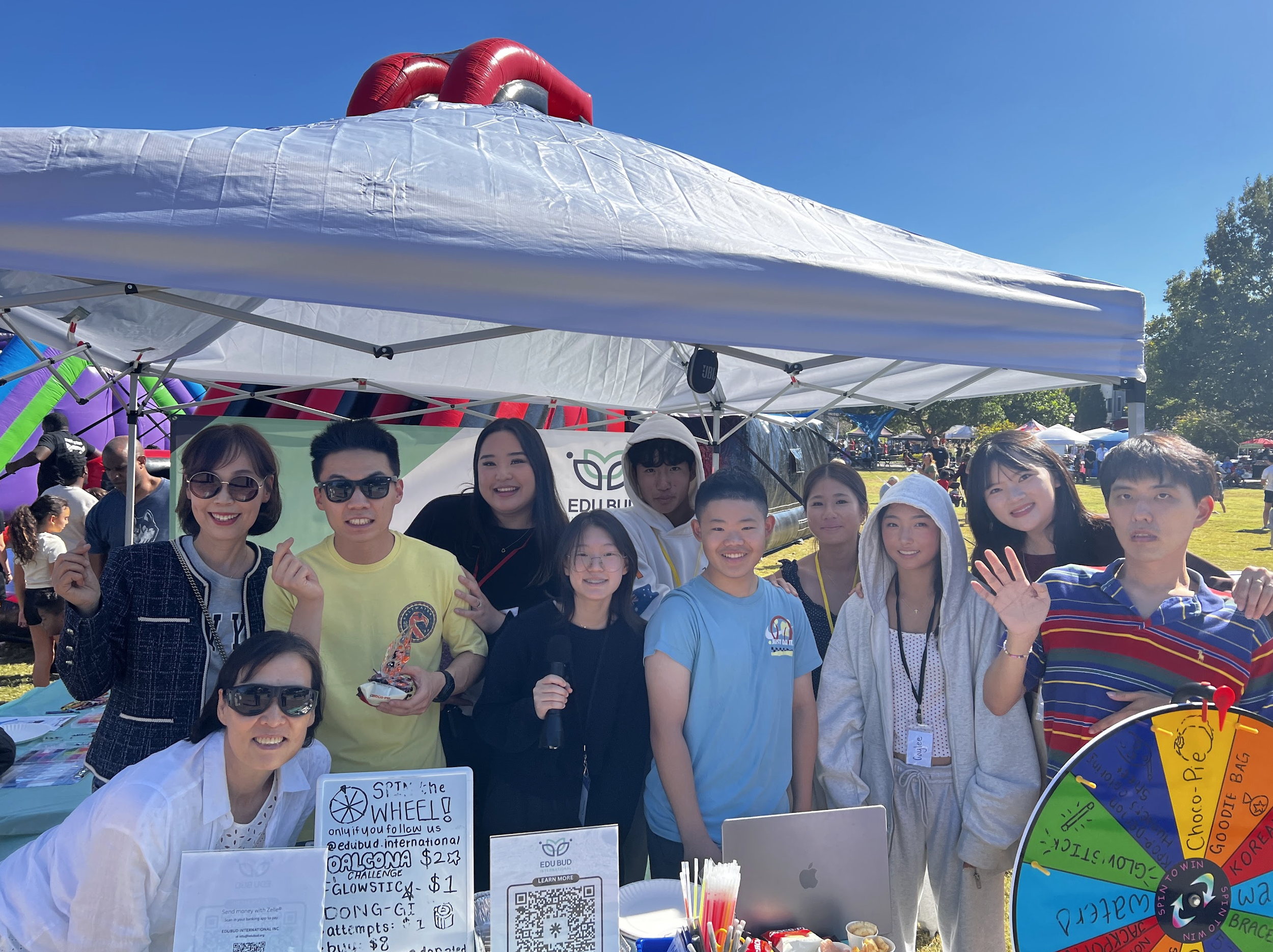 A group of people attending an outdoor event or fair, standing behind a booth with signs and colorful items, and smiling at the camera under a clear blue sky.