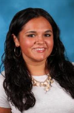Portrait of a woman with long, wavy black hair, wearing hoop earrings, a white top, and layered necklaces, smiling against a blue background.