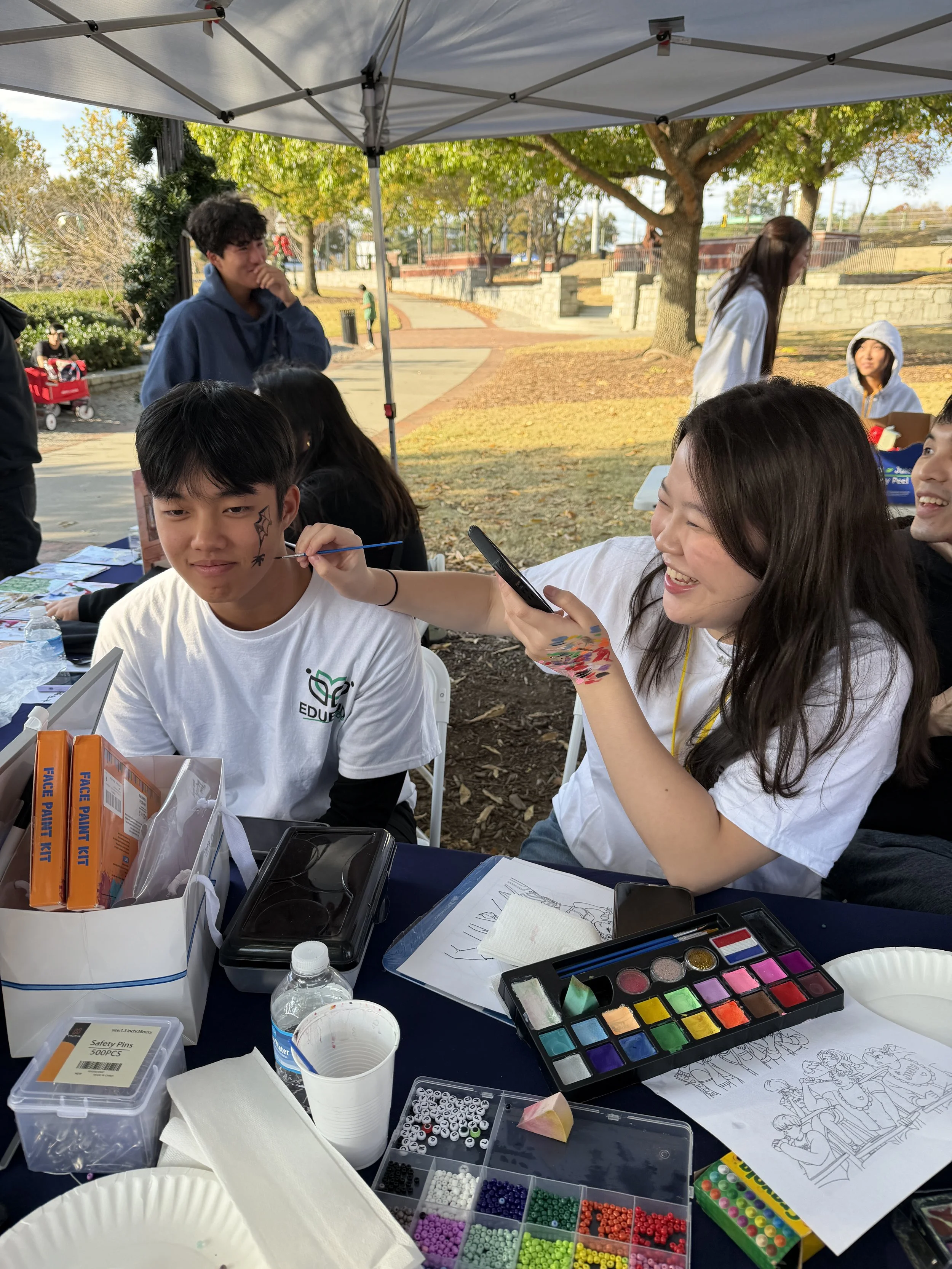 Group of young people at an outdoor arts and crafts table, with supplies like paint, beads, and coloring pages, under a canopy in a park with trees and a pathway in the background.