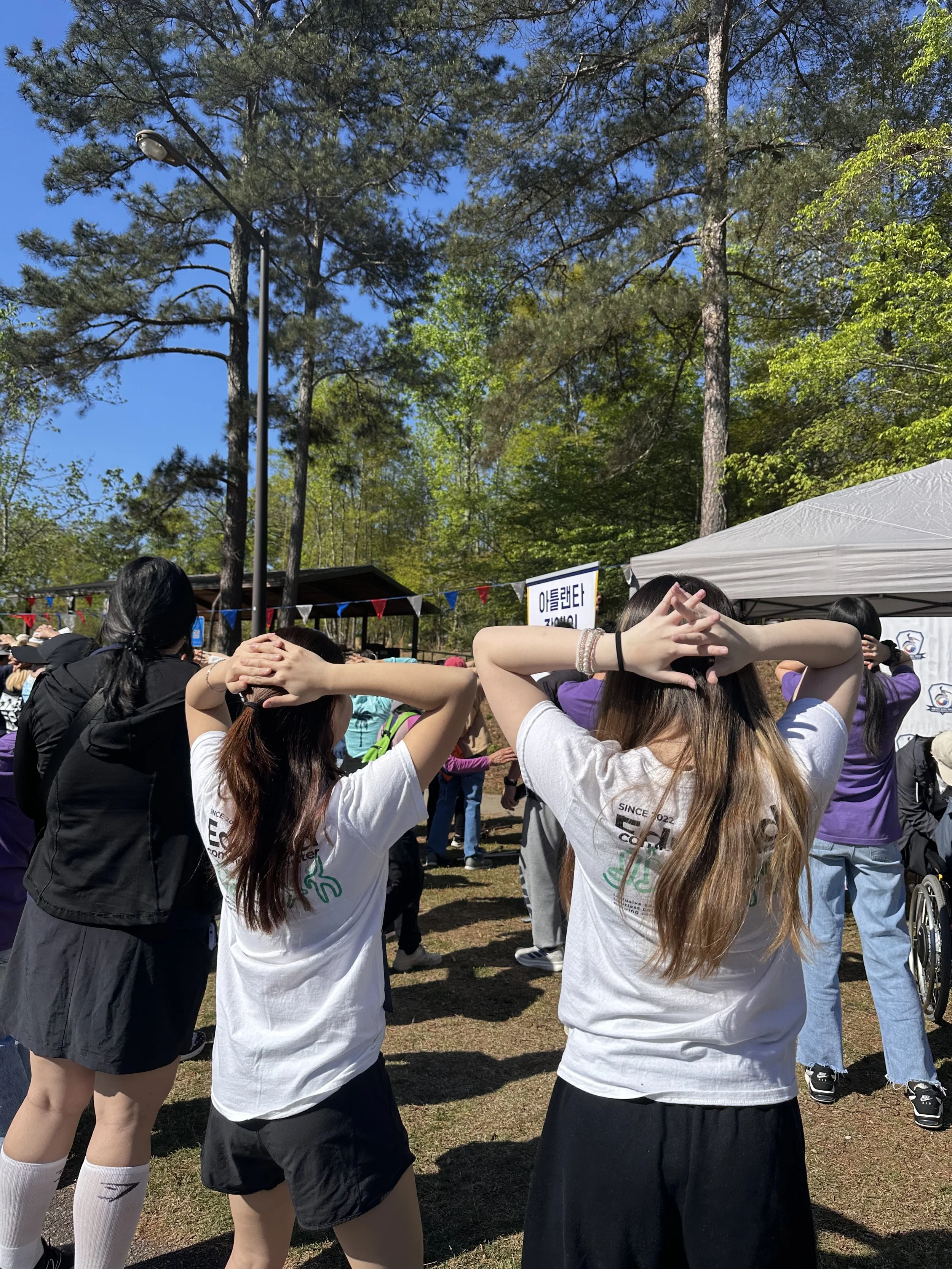 A group of young women participating in an outdoor event on a sunny day, with tall trees and a blue sky in the background. Some women have their hands on their heads, and they are dressed in casual athletic wear.
