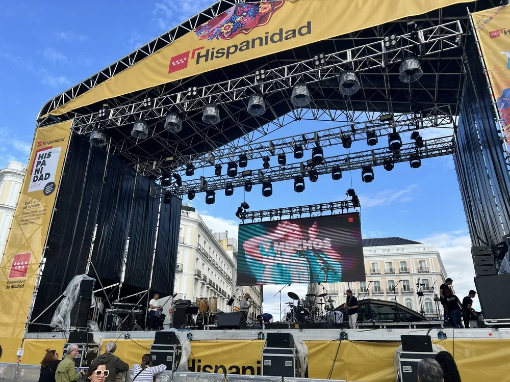 An outdoor stage with a yellow banner marked 'Hispanidad,' set up for a live music performance with musicians, equipment, and large screen, on a city street with white buildings and a blue sky.