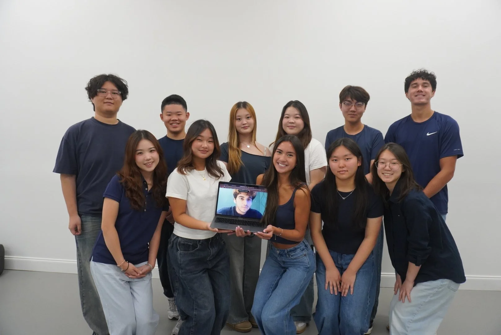Group of eleven young people standing against a plain white wall, some smiling and looking at the camera, two women holding a laptop displaying a young man's face.