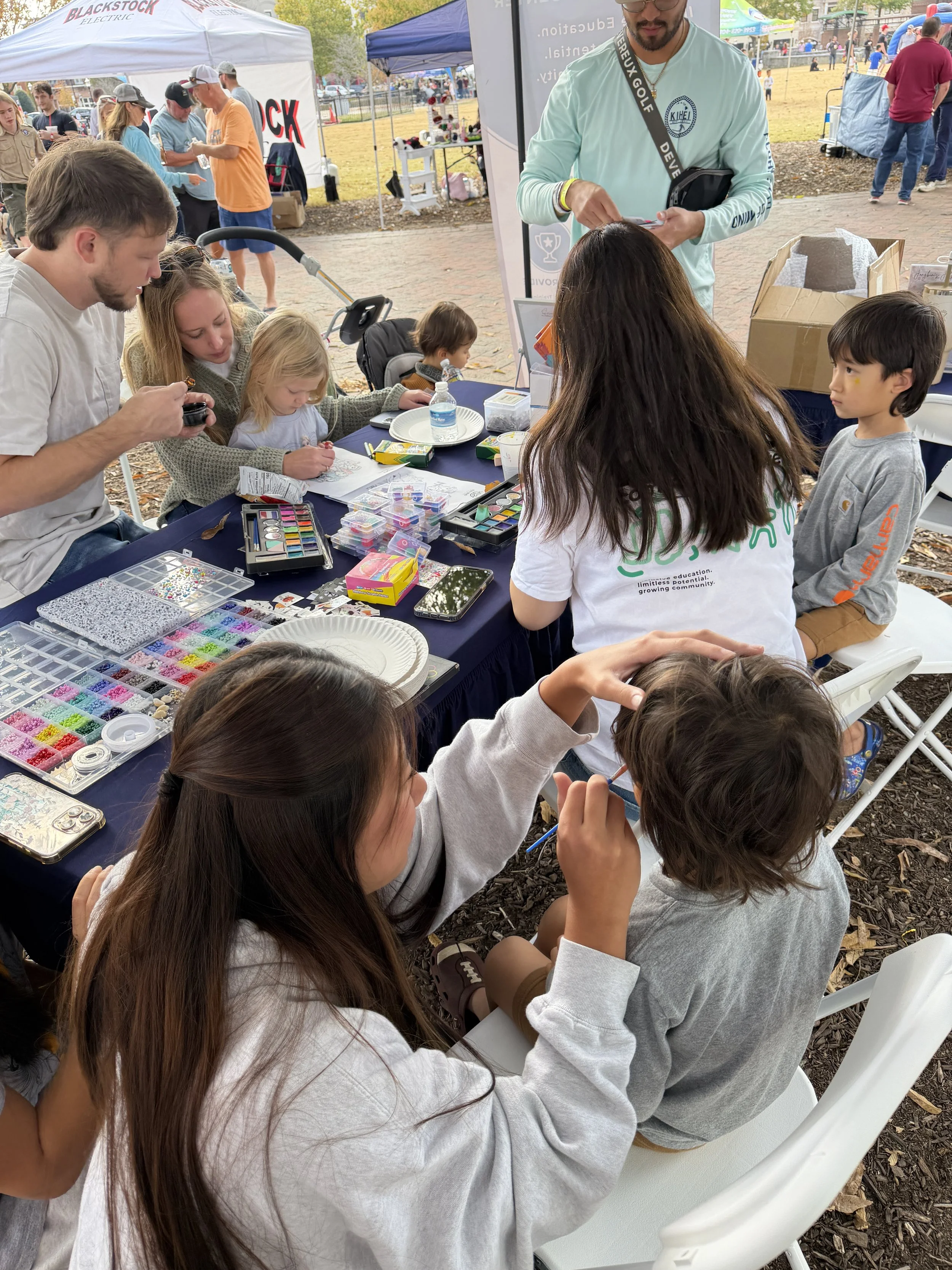 People at an outdoor craft event with tables filled with beads, jewelry making supplies, and children drawing or crafting. Tents and other event booths are visible in the background.