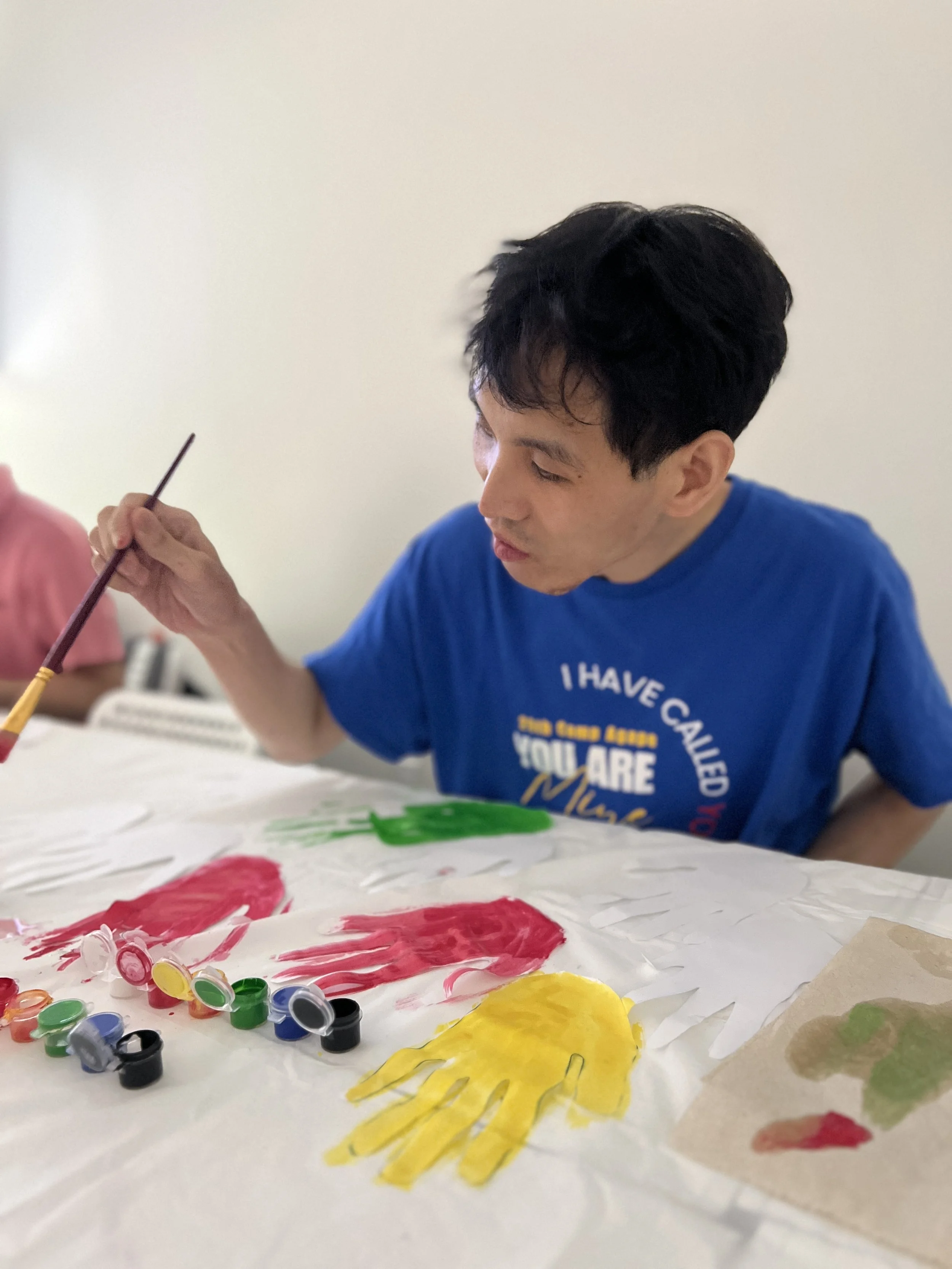 A man in a blue shirt is painting on a large piece of paper using various paints and brushes. The paper has colorful handprints and painted designs, including red, yellow, and green paint. The focus is on his side profile, and he appears to be engage