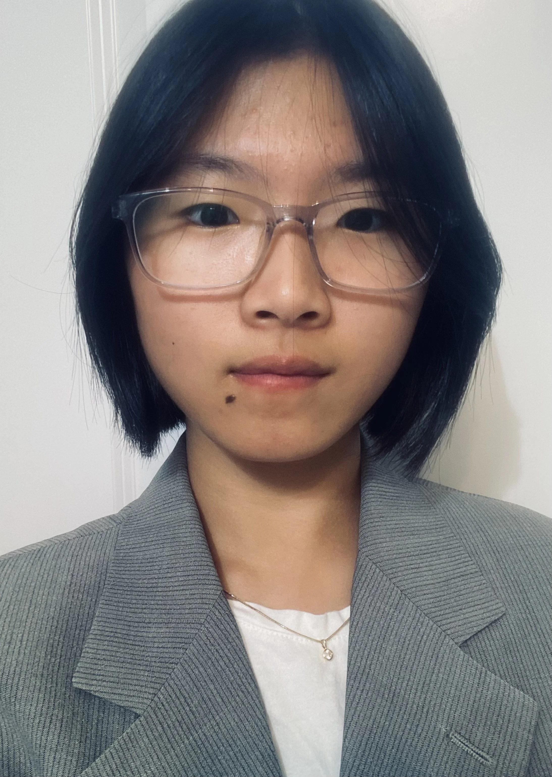 A woman with short black hair, wearing glasses, a gray blazer, a white shirt, and a gold necklace, standing against a plain wall.