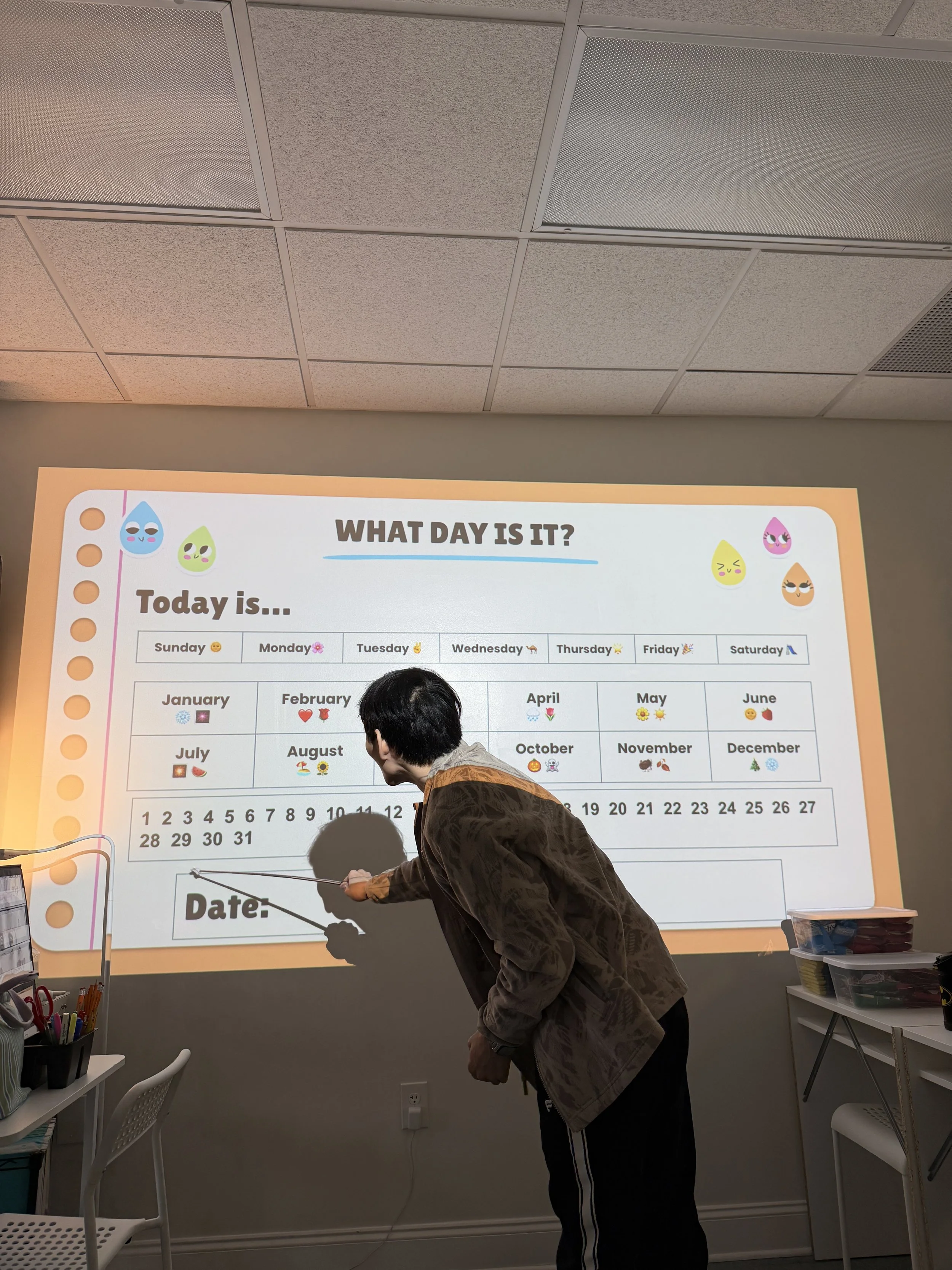 A person is pointing at a large projected calendar on a wall in a room that appears to be an office or classroom. The calendar features months, days, and emoji icons, with a colorful top border that has cute water droplet characters.