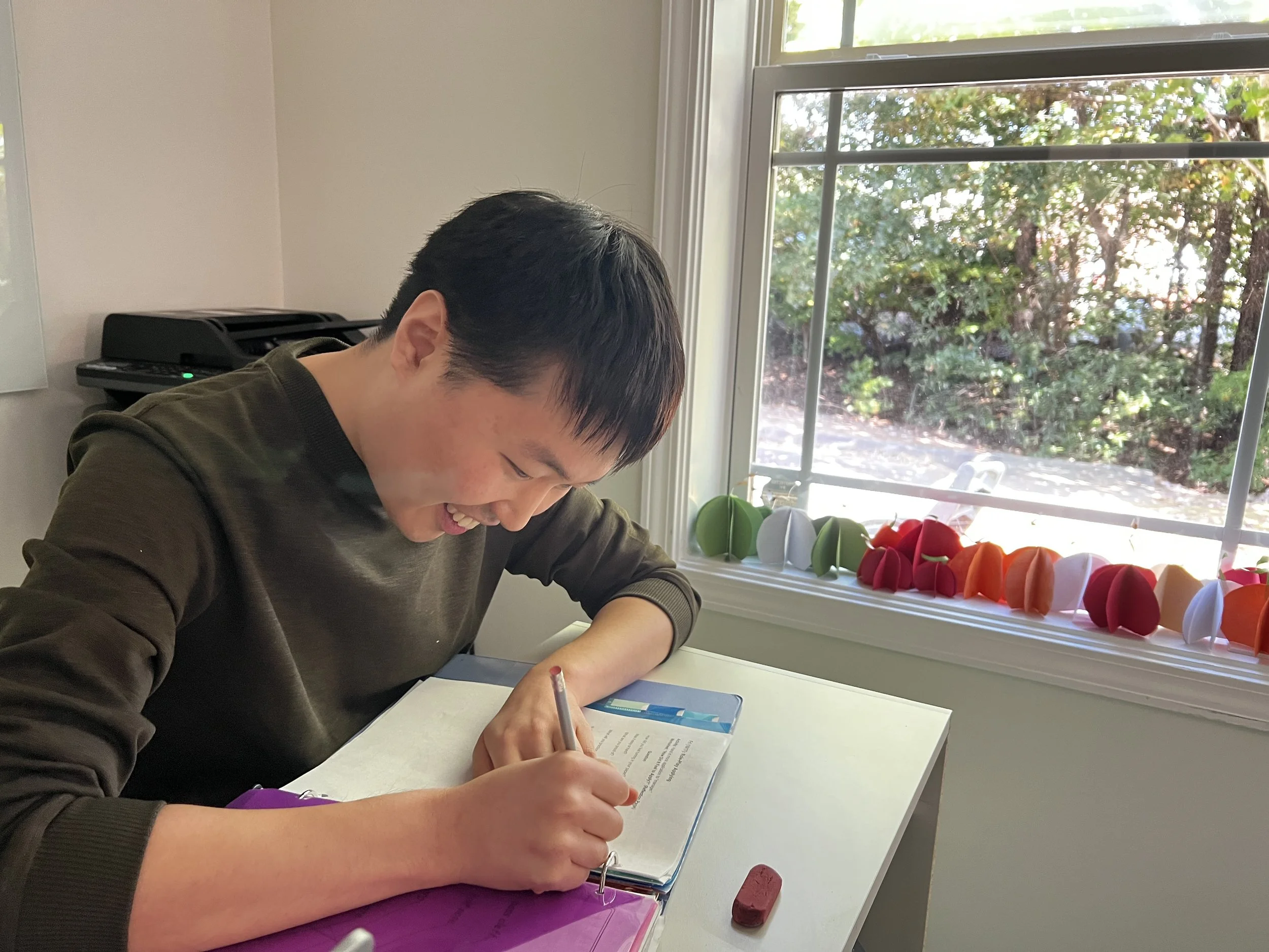 A person with short black hair sitting at a white desk, writing in a notebook, near a window with a view of green trees outside, with colorful paper pumpkins on the windowsill.