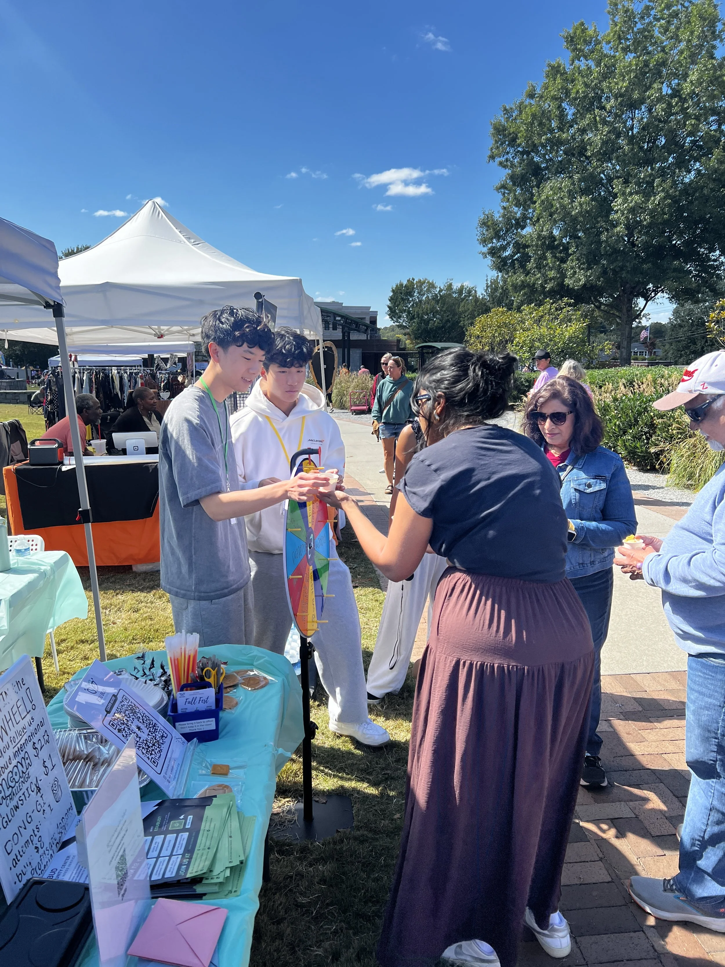 People at an outdoor event engaging in conversations near a table with pamphlets and a game wheel. Tents and other people are visible in the background, with a clear blue sky and trees surrounding the area.