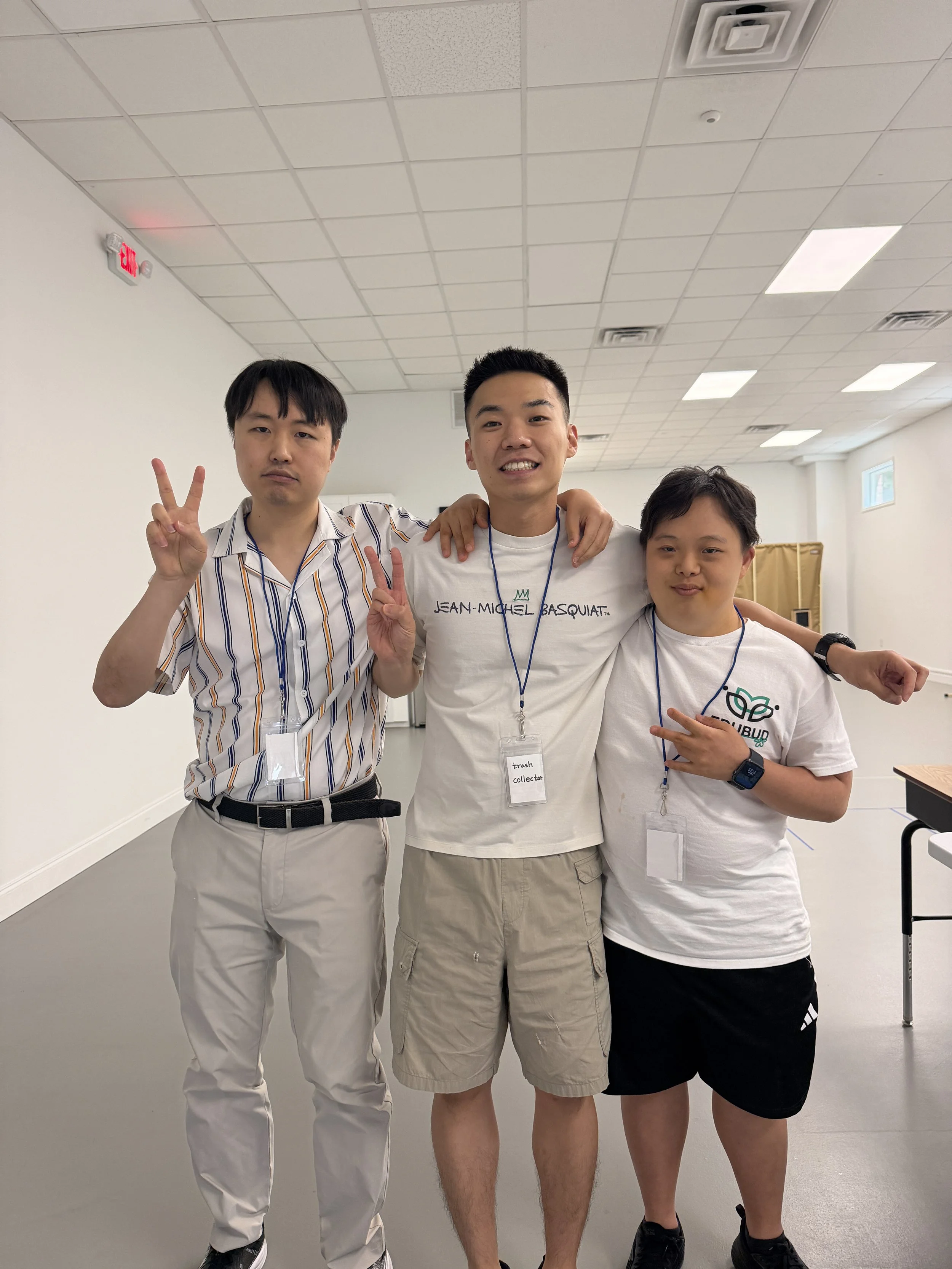 Three young men standing together indoors, smiling and making peace signs. The man in the middle is wearing a white t-shirt with text, beige shorts, and a lanyard with a badge. The man on the left is in a striped shirt and light-colored pants, while 