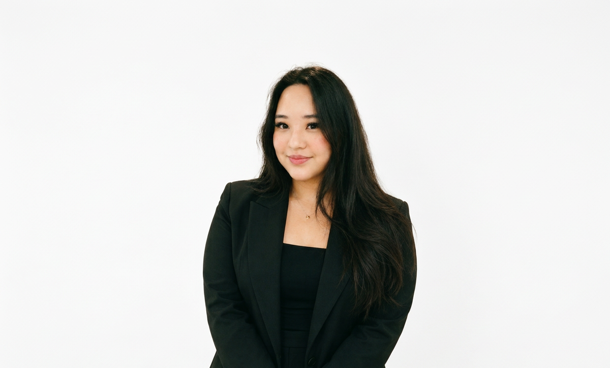 A woman with long dark hair in a black blazer and top, standing against a plain white background, smiling slightly.