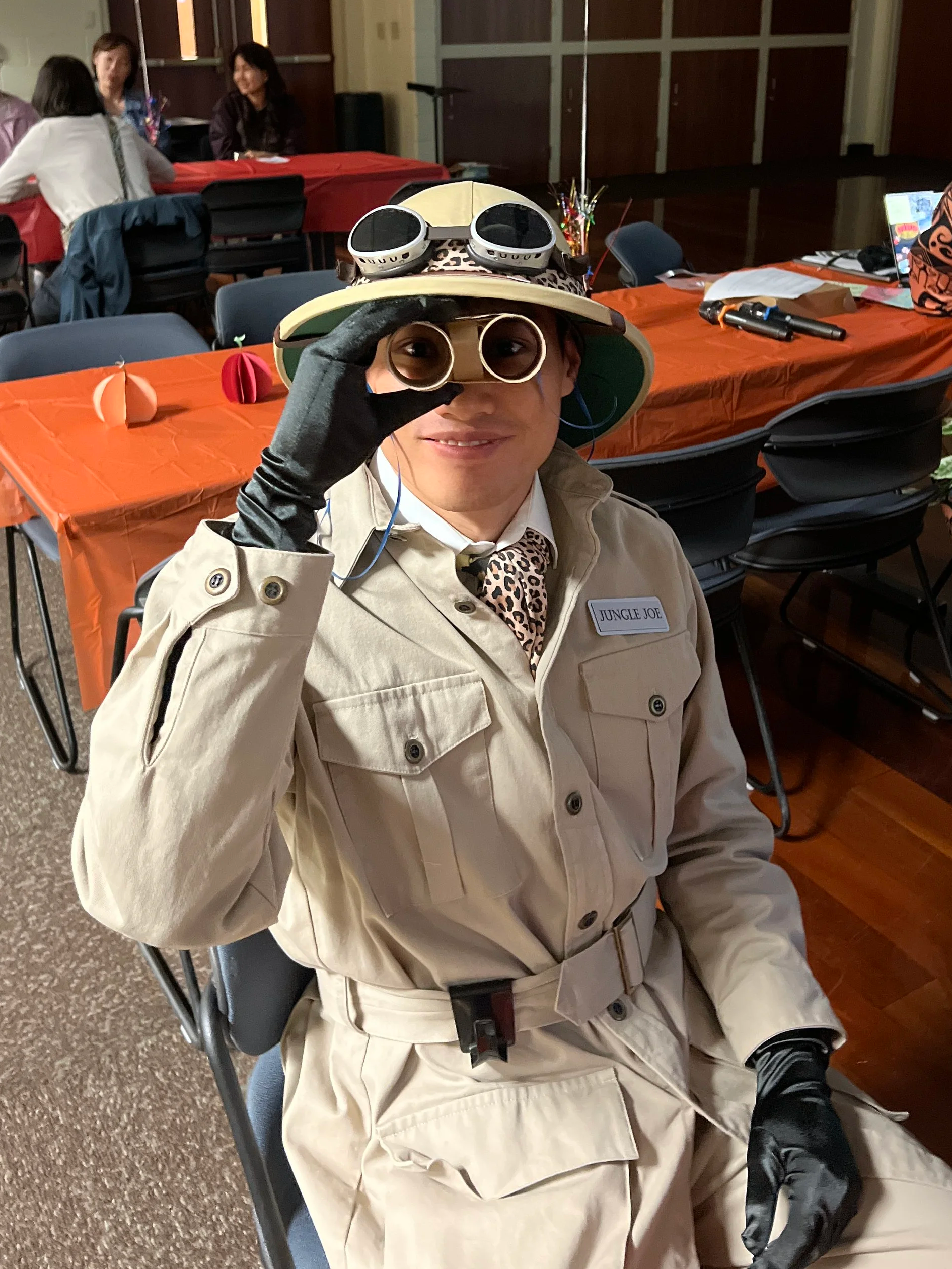 Person dressed as a jungle explorer wearing a beige safari outfit, black gloves, sunglasses, and a wide-brimmed hat with goggles, sitting in a room with decorated tables and other people.