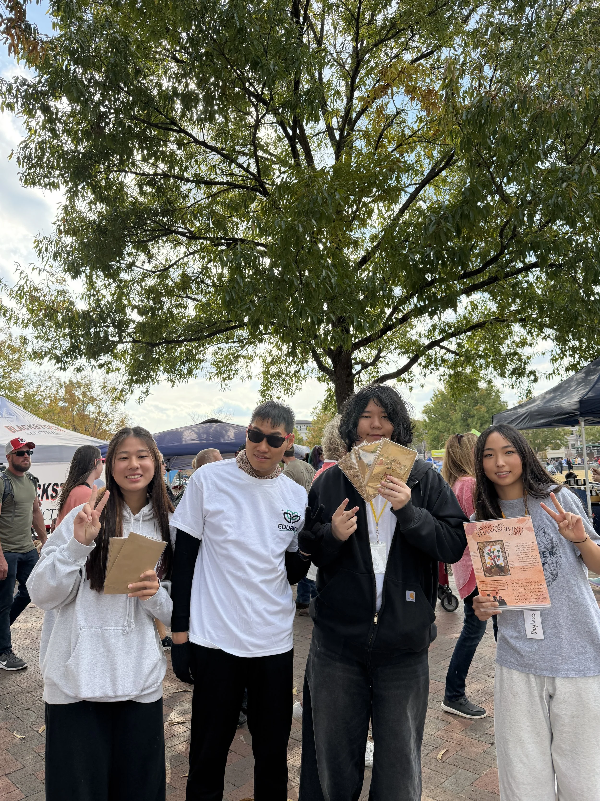 Group of young people standing outdoors under a large tree at a festival or fair, holding pamphlets and making peace signs, with tents and other people in the background.