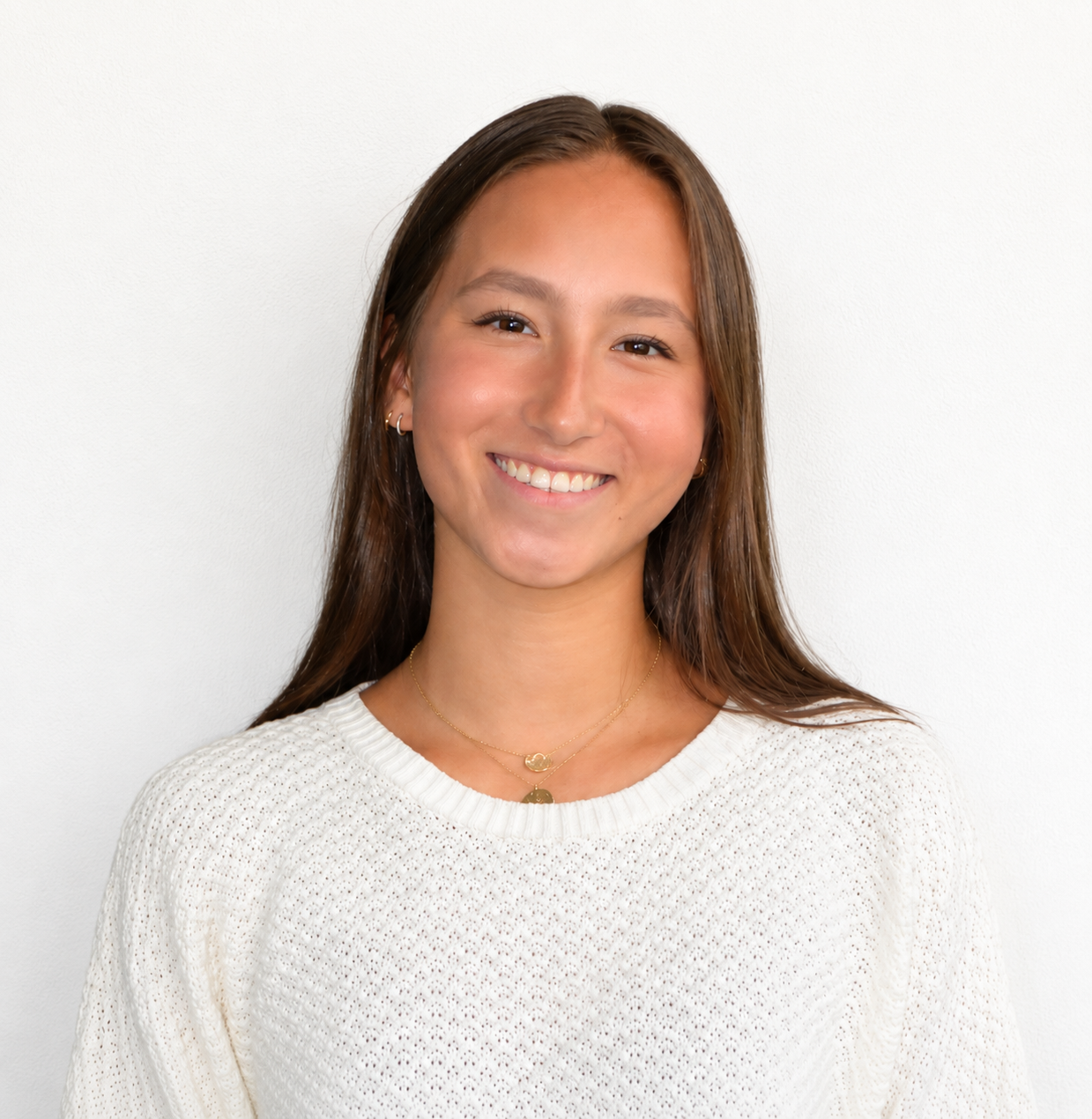 A young woman with long brown hair smiling, wearing a white textured sweater and gold jewelry, standing against a plain white wall.