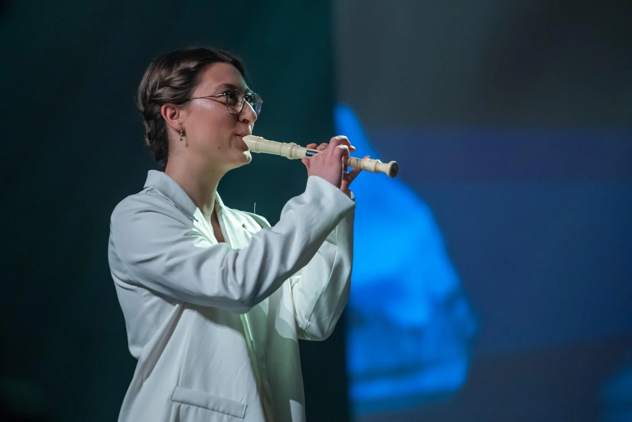 Marianne Paquin joue de la flûte à bec sur scène, portant un blouson blanc. Éclairage bleu en arrière-plan.