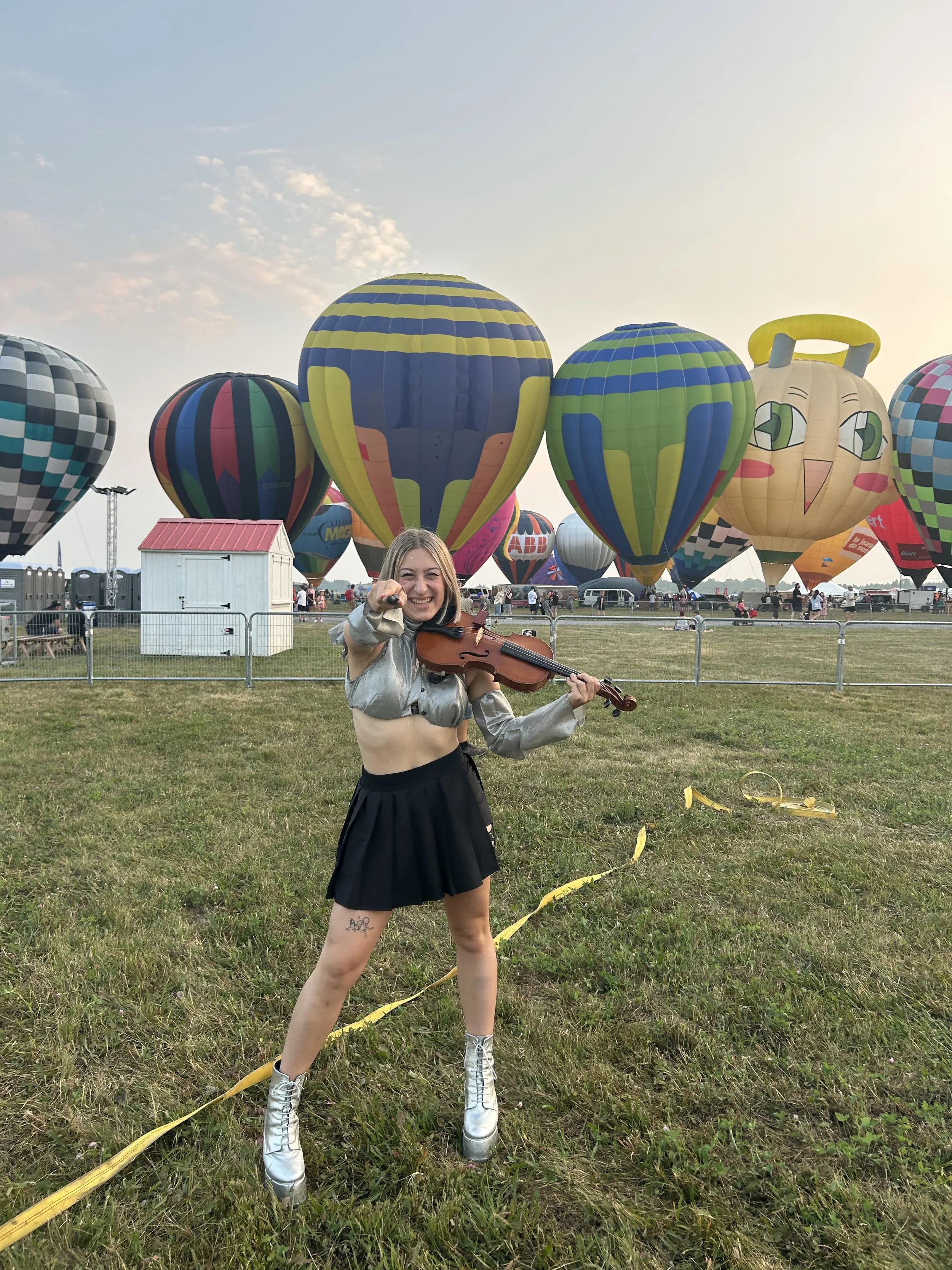 Marianne Paquin dans un champ d'envolée de montgolfières au coucher de soleil. Vêtue d'argent, d'une mini-jupe noire, elle tient un violon et pointe son archet vers la caméra.