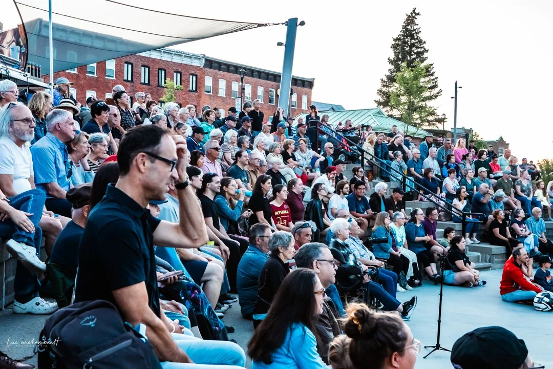 Une foule de personnes assises en plein air lors d'un spectacle de Marianne Paquin, avec un décor urbain en arrière-plan.
