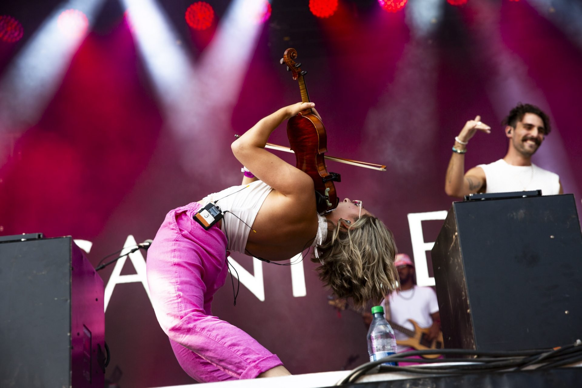 Marianne Paquin joue du violon sur scène à Osheaga avec un public et des musiciens en arrière-plan, sous des lumières colorées. Elle pose penchée vers l'arrière.