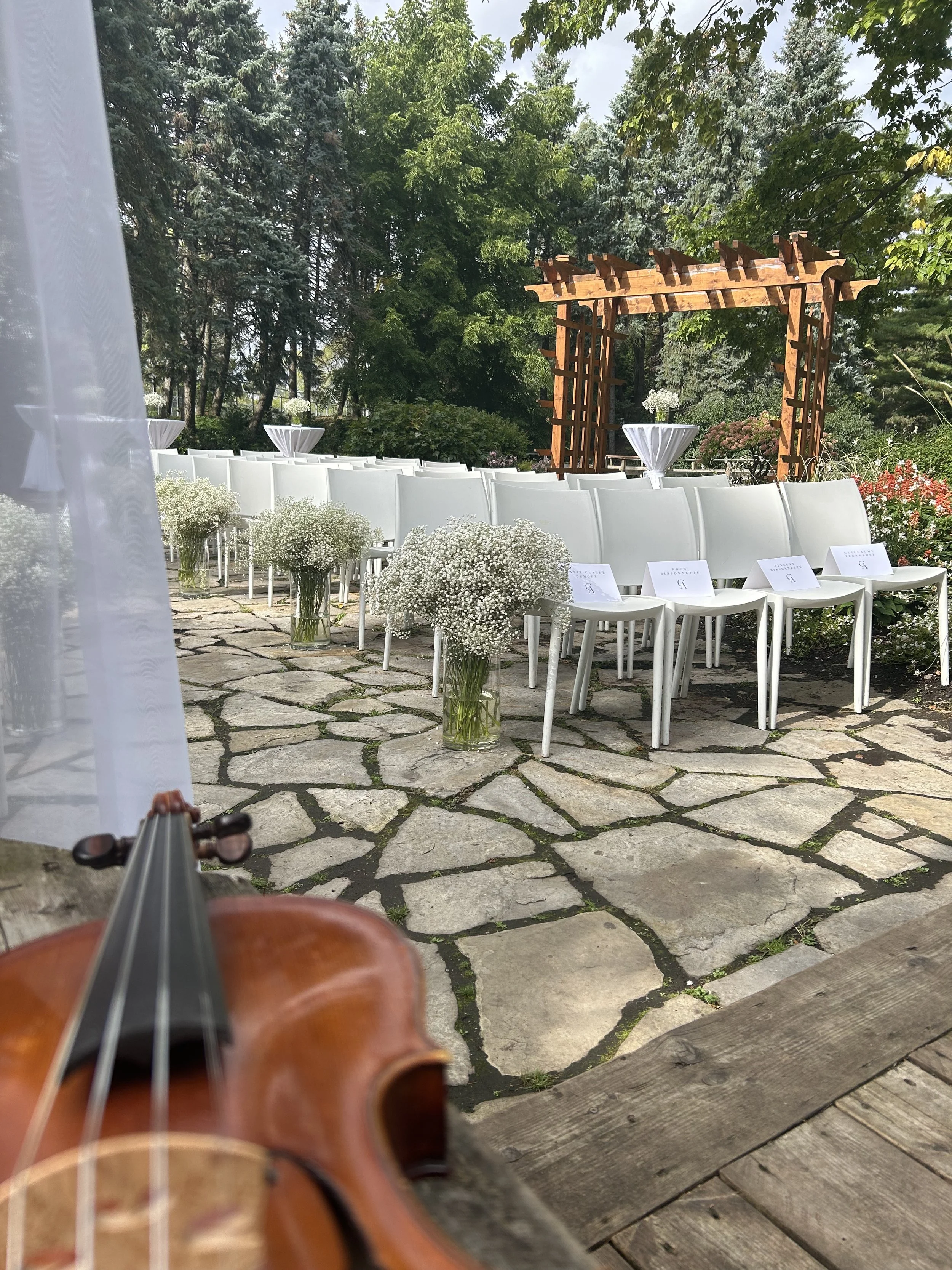 Une scène en plein air avec des chaises blanches, des tables décorées de fleurs blanches, une arche en bois et un fond d'arbres verts, suggérant un mariage. Un violon en bas à droite.