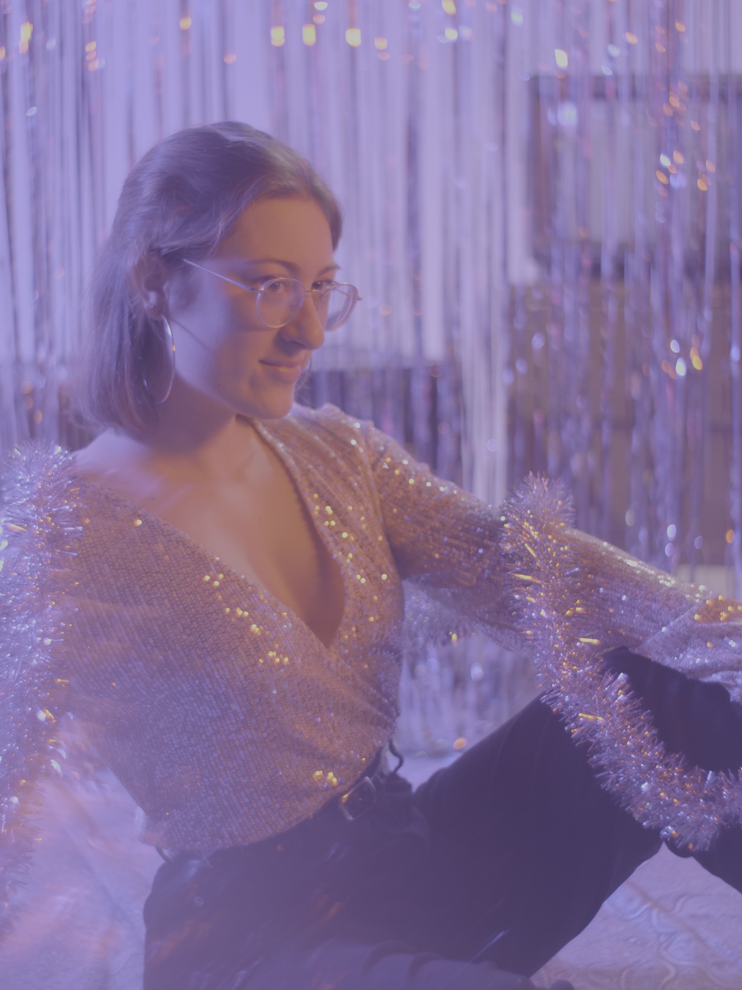 Marianne Paquin avec des lunettes, portant un haut scintillante dorée, assise dans un décor festif avec des guirlandes et une ambiance lumineuse violette et argentée.