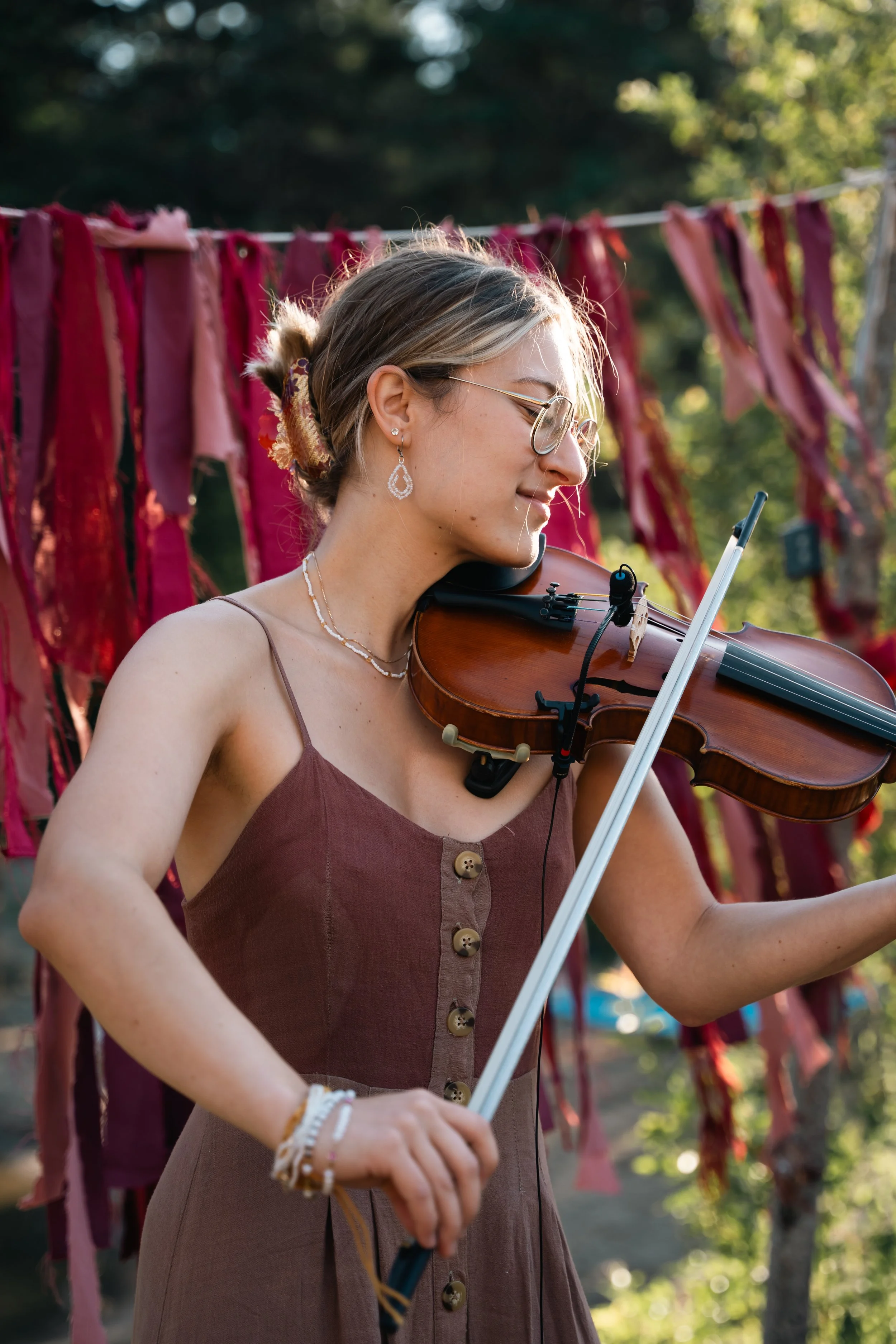 Marianne Paquin jouant du violon en extérieur, portant des lunettes, des bijoux, et une robe marron à boutons, avec des morceaux de tissu suspendus en arrière-plan.