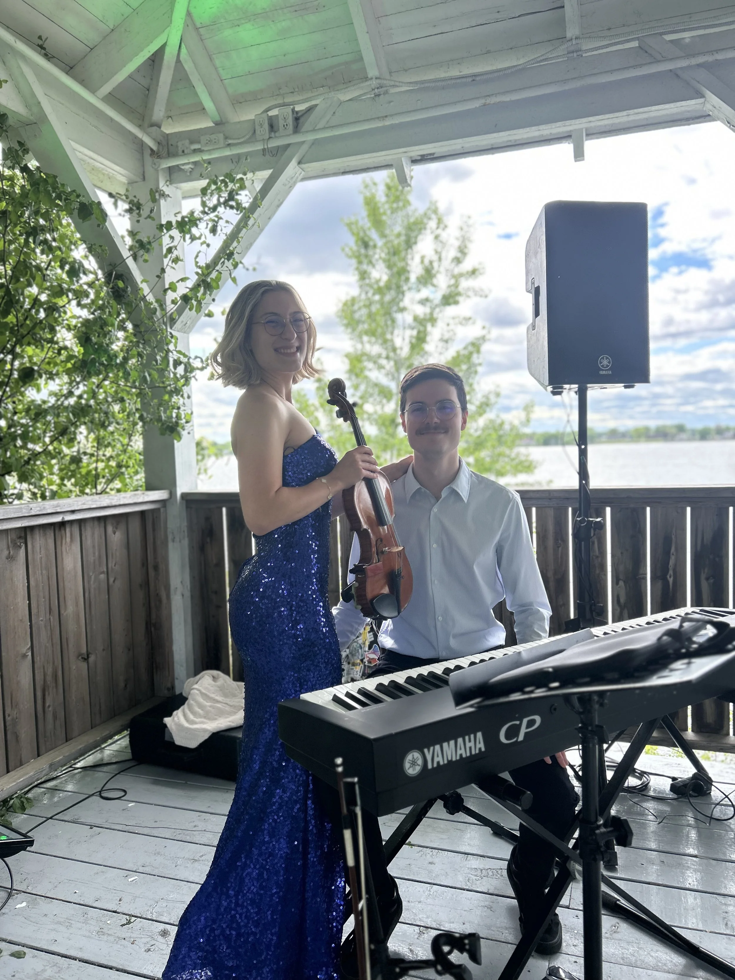 Marianne Paquin et Samuel Therrien jouent lors d'une performance en plein air. Marianne en robe bleue à sequins tient un violon, tandis qu'e Samuel assis joue du piano numérique sous un toit en bois, avec un haut-parleur à côté.