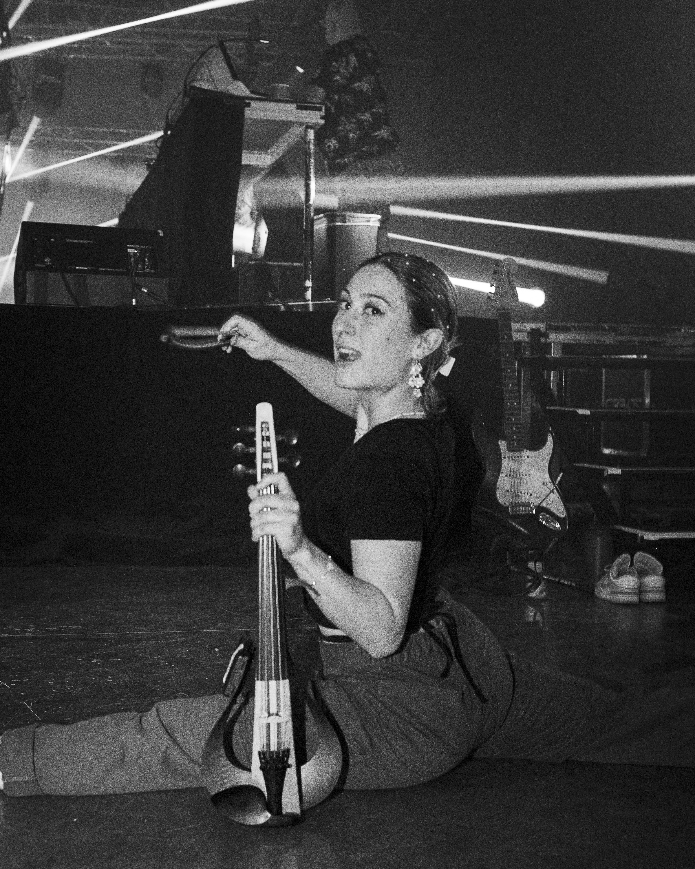 Image en noir et blanc de Marianne Paquin en position de split. Elle tient un violon électrique et pointe la caméra de son archet. Pendant un spectacle de musique électronique.