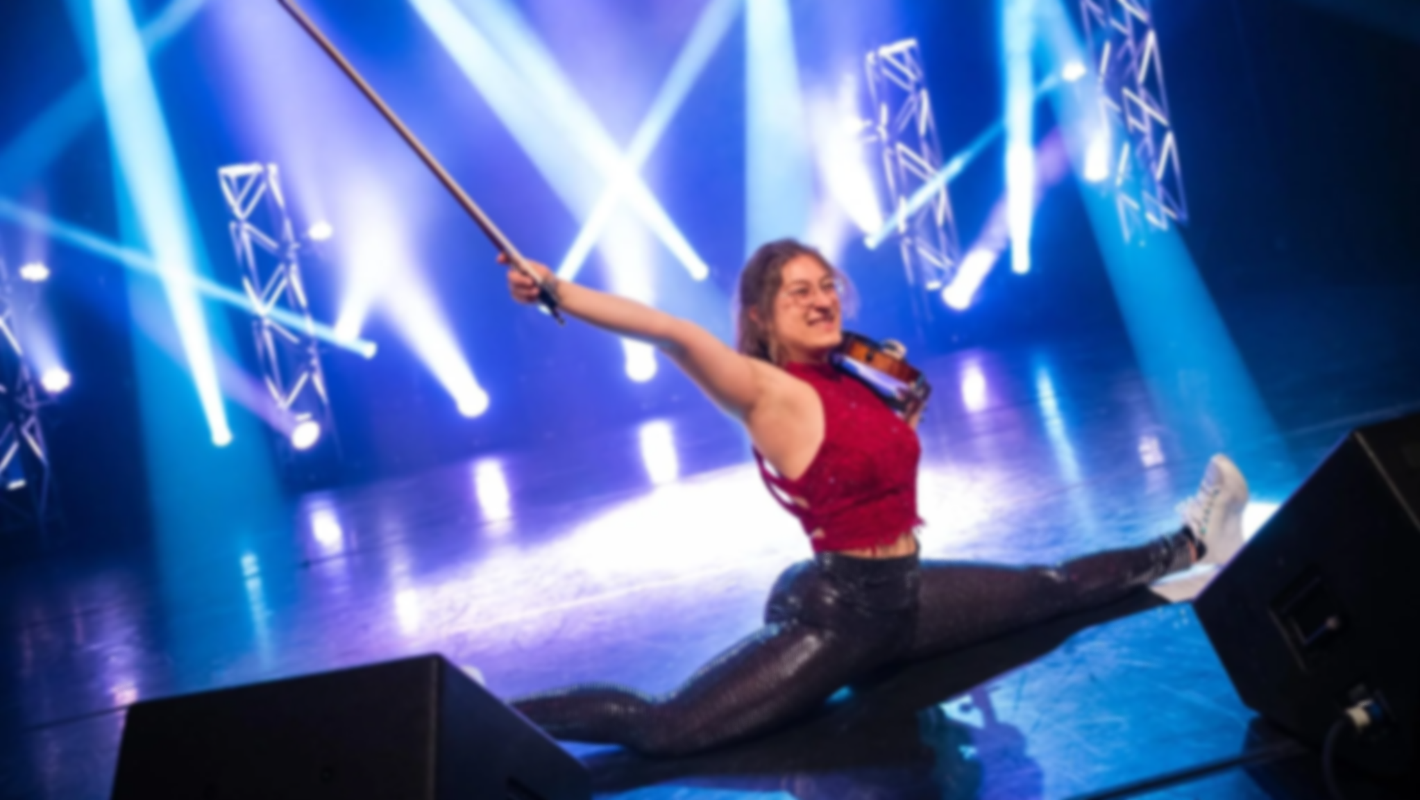 Marianne Paquin jouant du violon sur scène, en position de split, sous un éclairage de spectacle avec des faisceaux de lumière bleus et blancs.