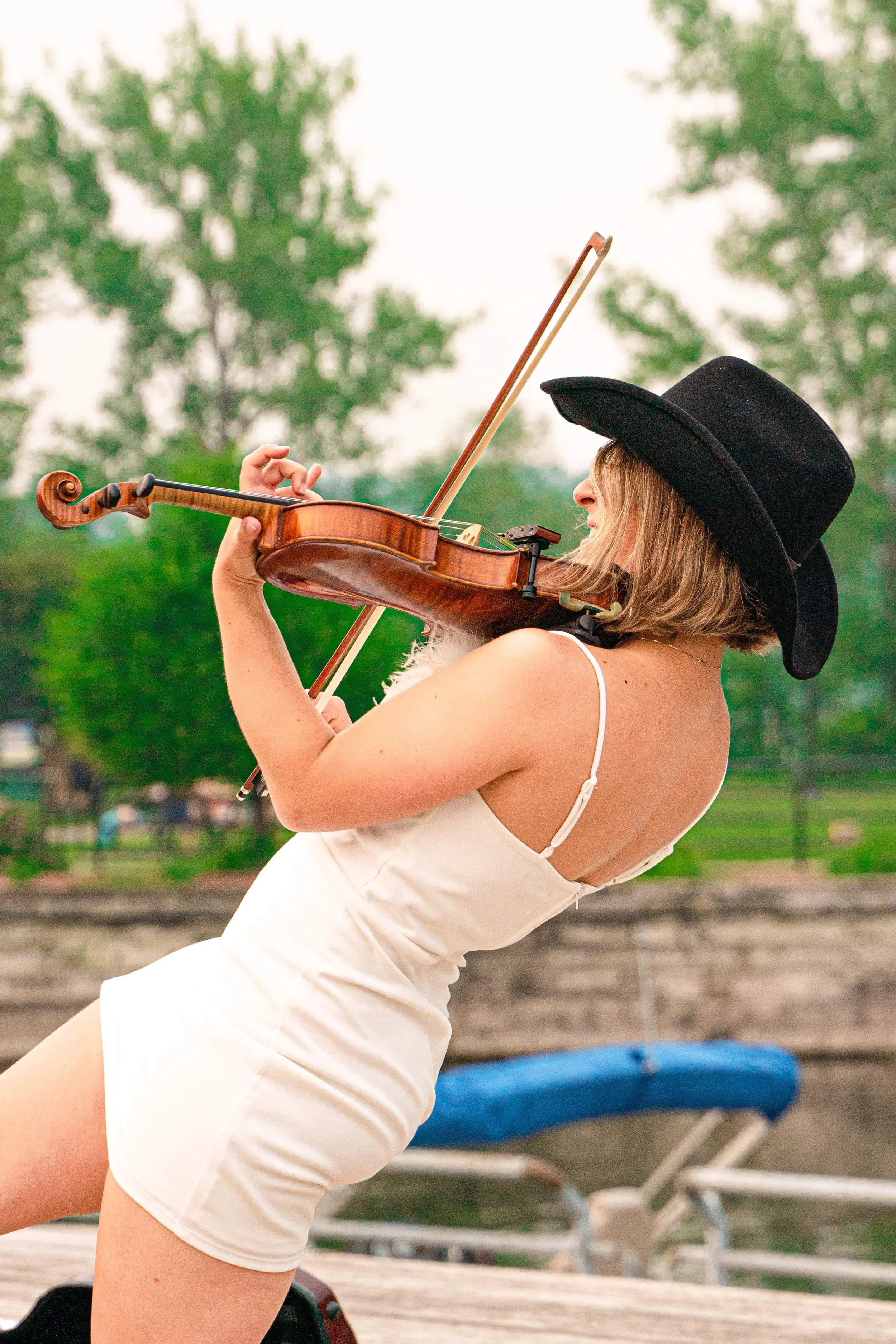 Marianne Paquin joue du violon en extérieur, portant un chapeau de cowboy noir et une petite robe blanche, avec des arbres verts en arrière-plan.