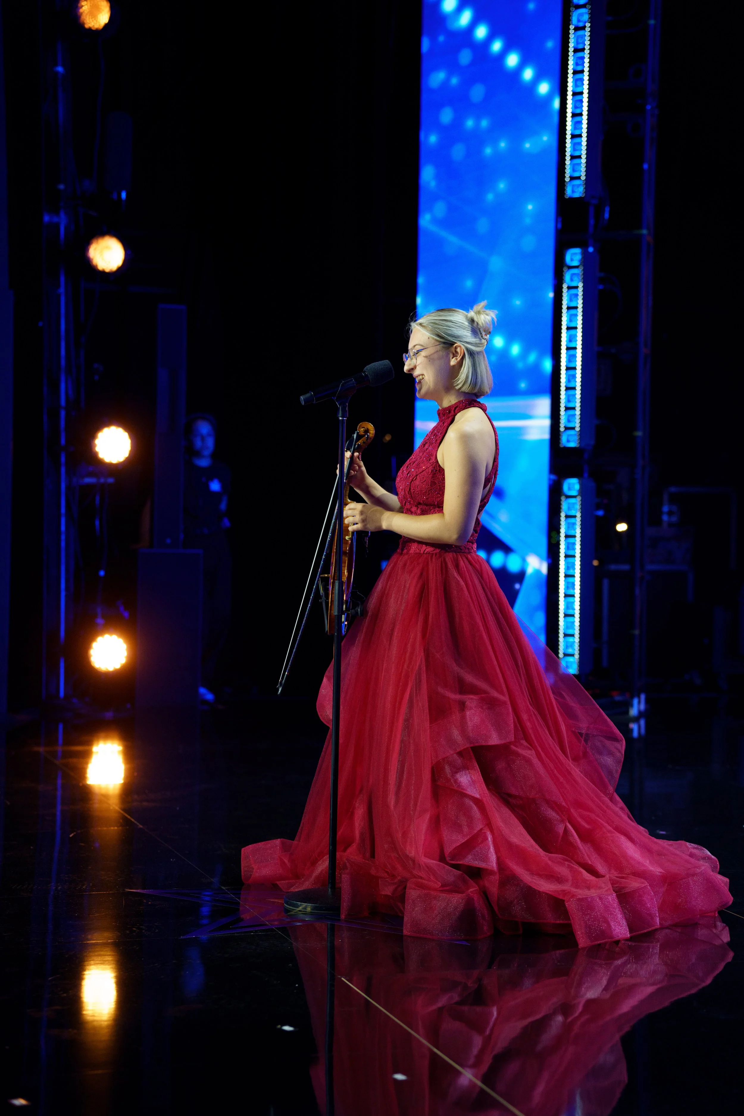 Marianne Paquin en longue robe de soirée rouge, tenant un violon, parle dans un micro sur une scène avec des lumières bleues lors de son audition pour "Quel Talent!".
