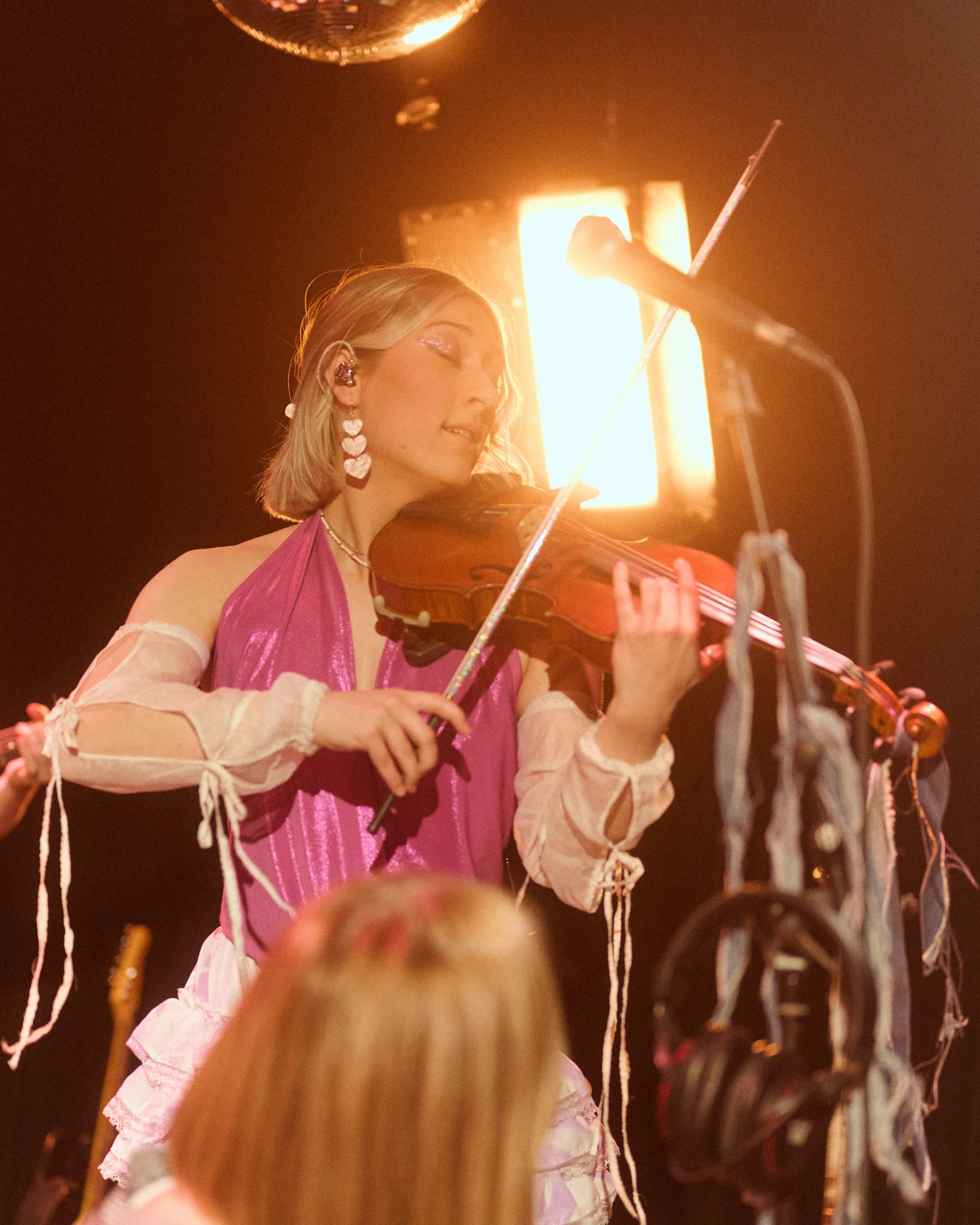 Marianne Paquin jouant du violon lors d'une performance, avec une lumière intense derrière elle, portant une robe rose. Décorations de rubans autour d'elle.