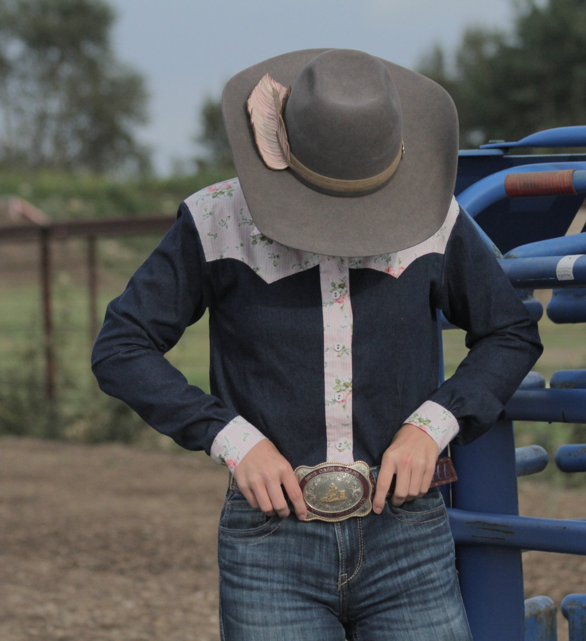 Person in western attire, wearing a cowboy hat with a feather, a patterned western shirt, and jeans, standing outdoors near a blue fence.
