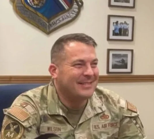 A man in military uniform smiling and sitting at a desk with framed photos and a military emblem on the wall behind him.
