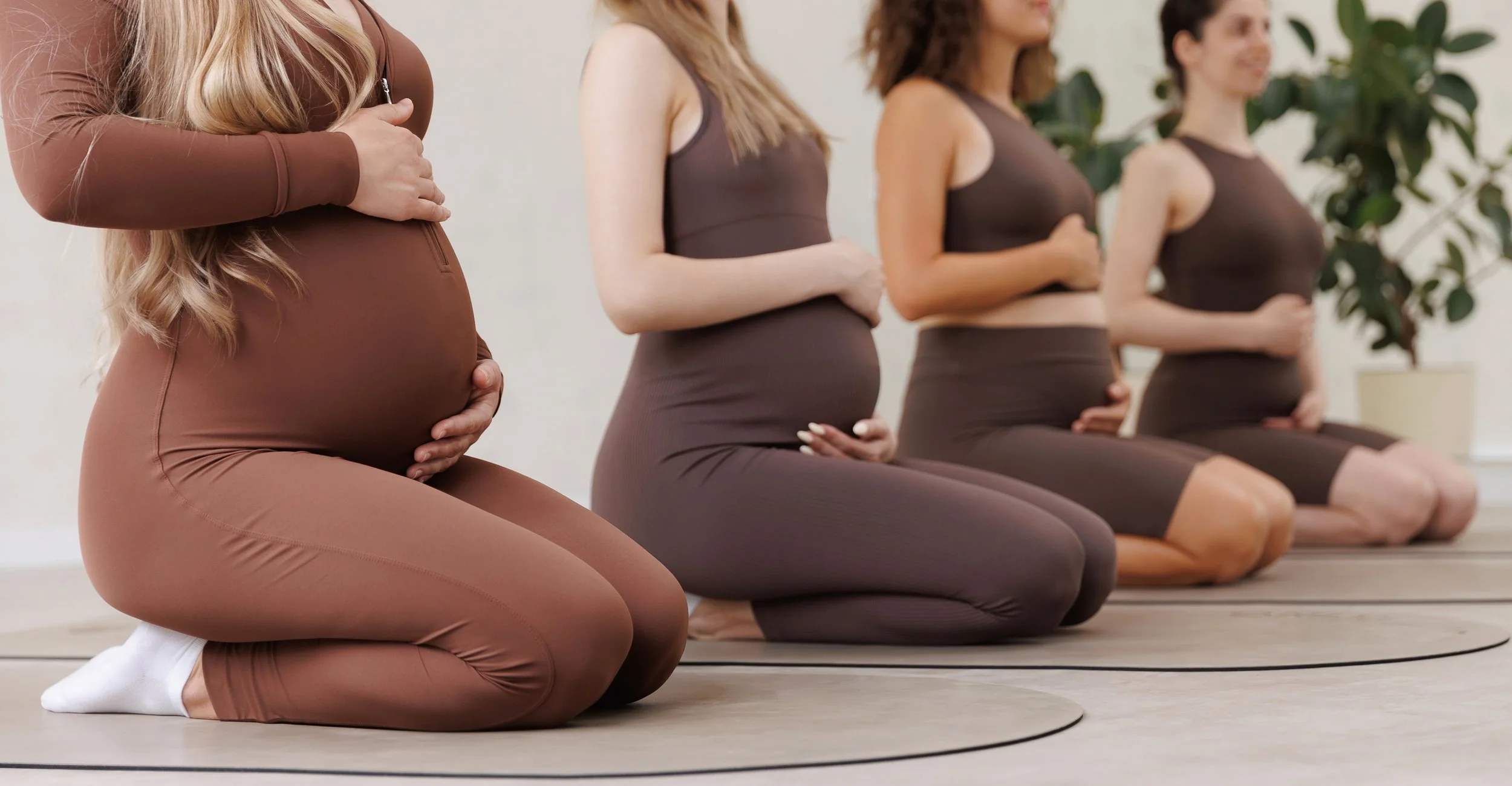 Four women practicing yoga in a kneeling position, holding their bellies with their hands, during a class held in a well-lit room with plants.