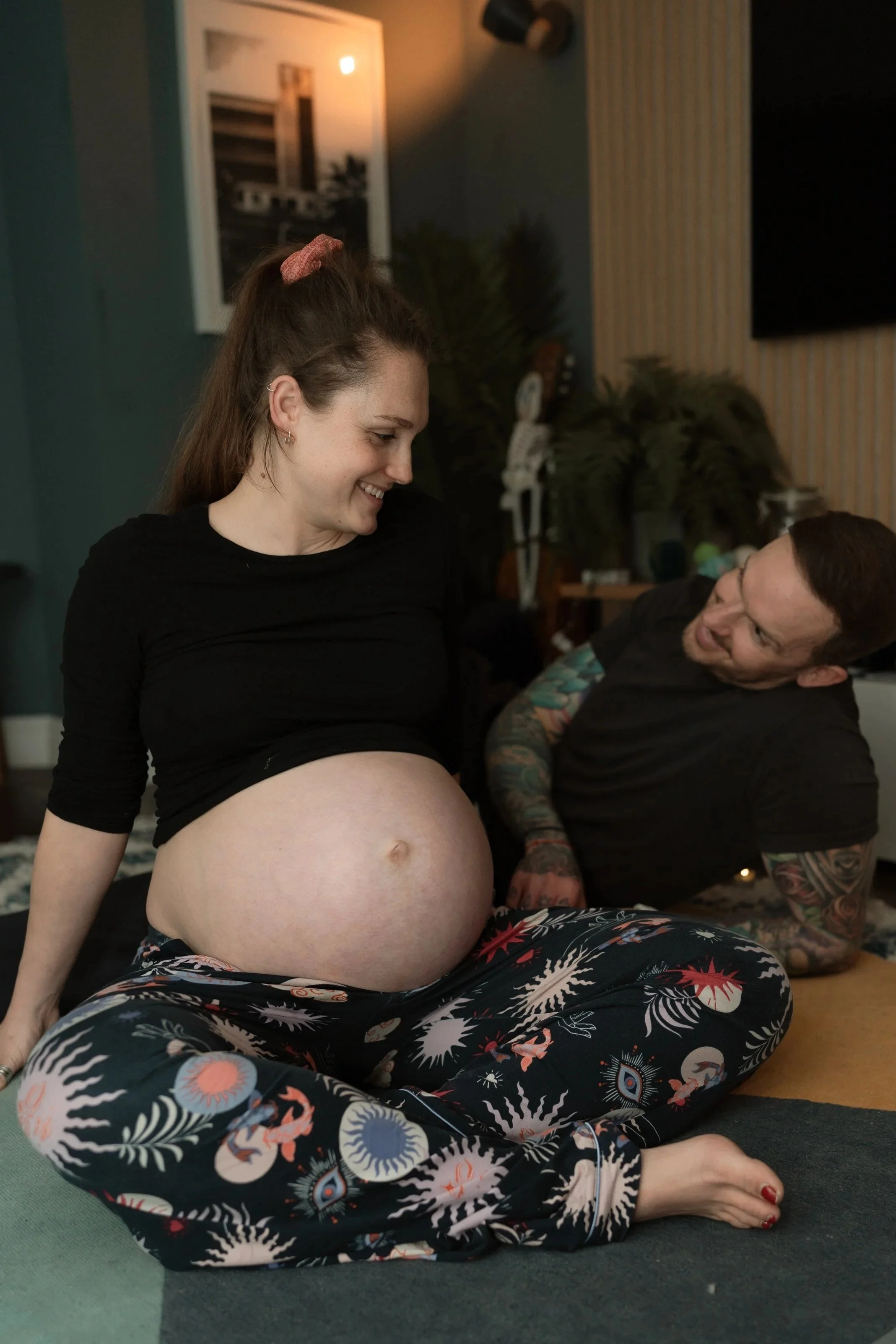 Pregnant woman sitting cross-legged on the floor, smiling, with man behind her touching her belly, in a cozy room with furniture and plants.