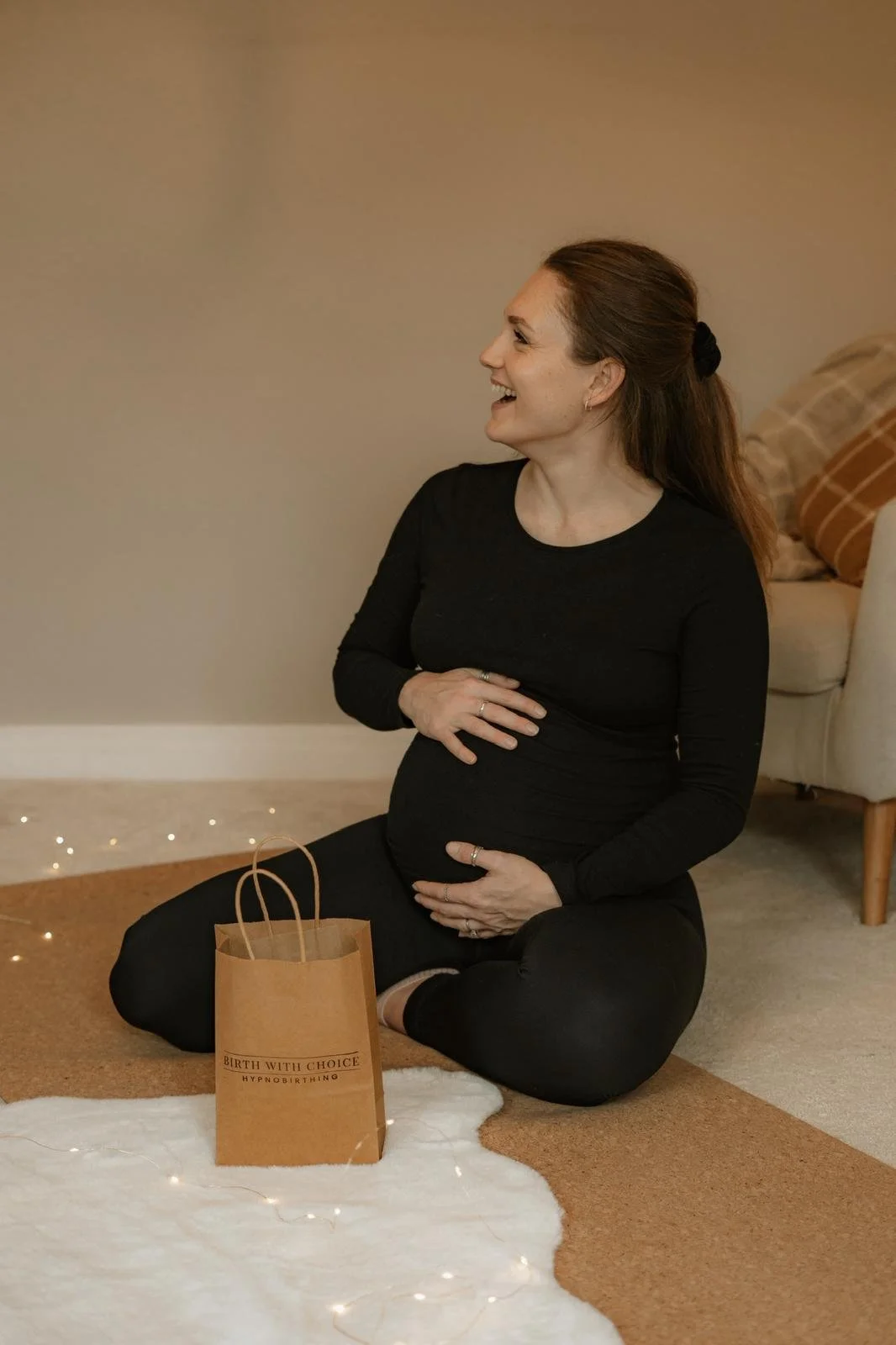 A smiling pregnant woman sitting on the floor with her hands on her belly, wearing a black long-sleeve shirt and black pants. There is a small brown gift bag with the text "Birth with Choice Hypnobirthing" on it, placed in front of her on a white fur rug, with a beige and white room background and some string lights.