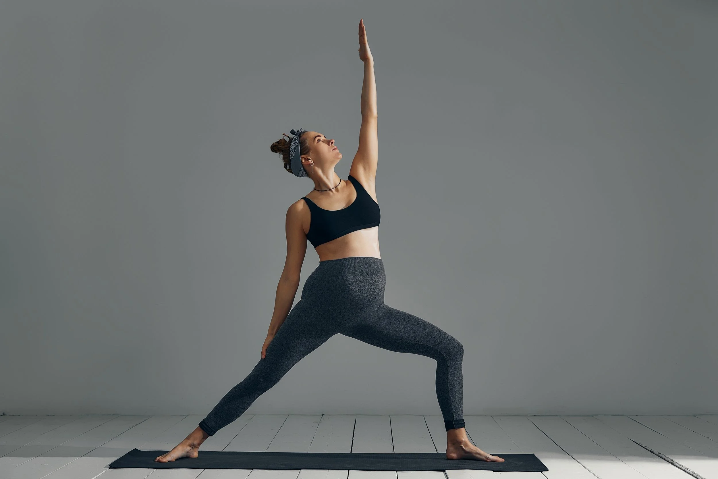 A woman practicing yoga indoors, performing a Warrior pose on a black yoga mat, wearing a black sports bra, gray leggings, and a headband.