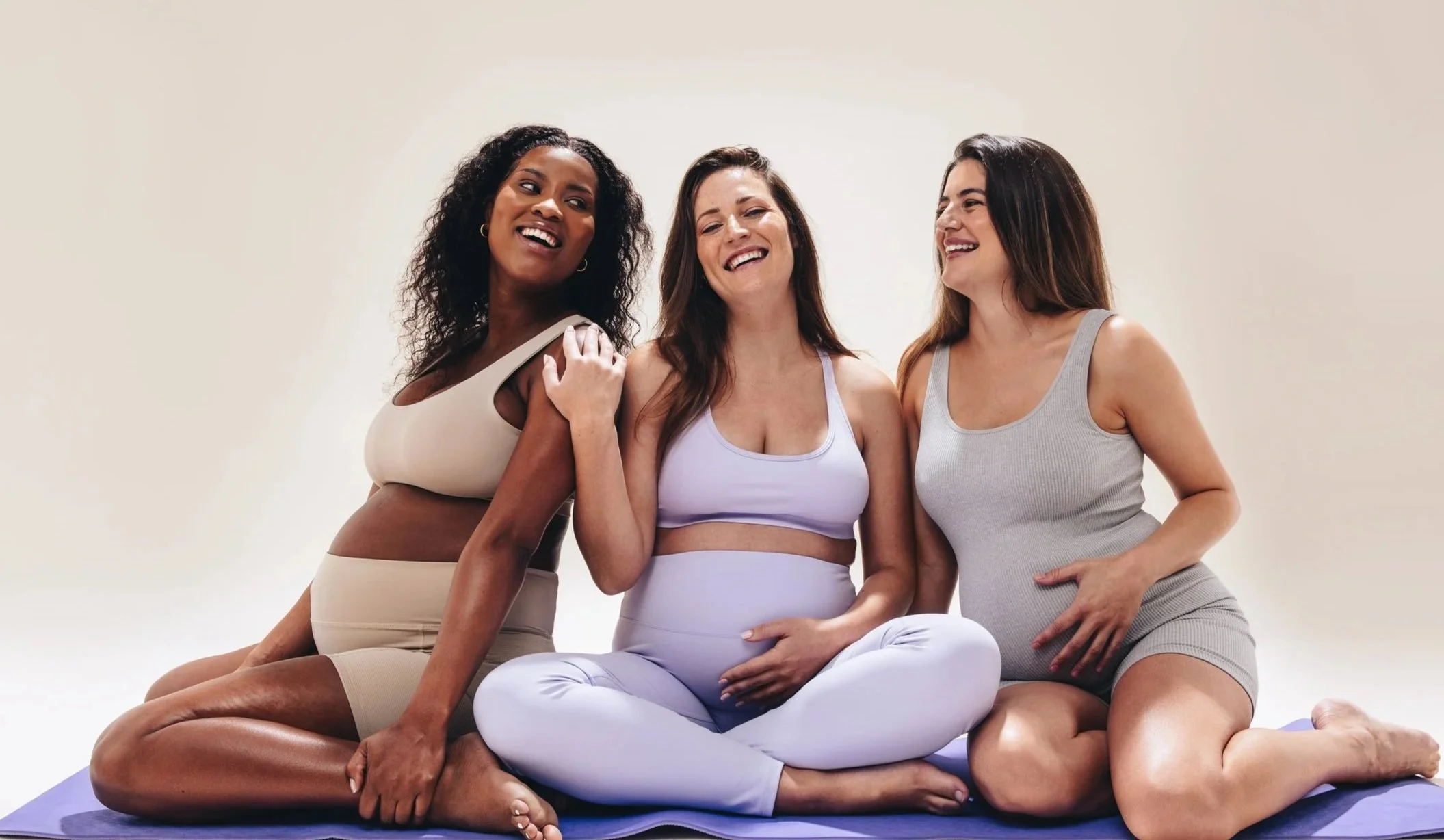 Three women are sitting on a yoga mat, smiling and laughing together, dressed in comfortable athletic wear.