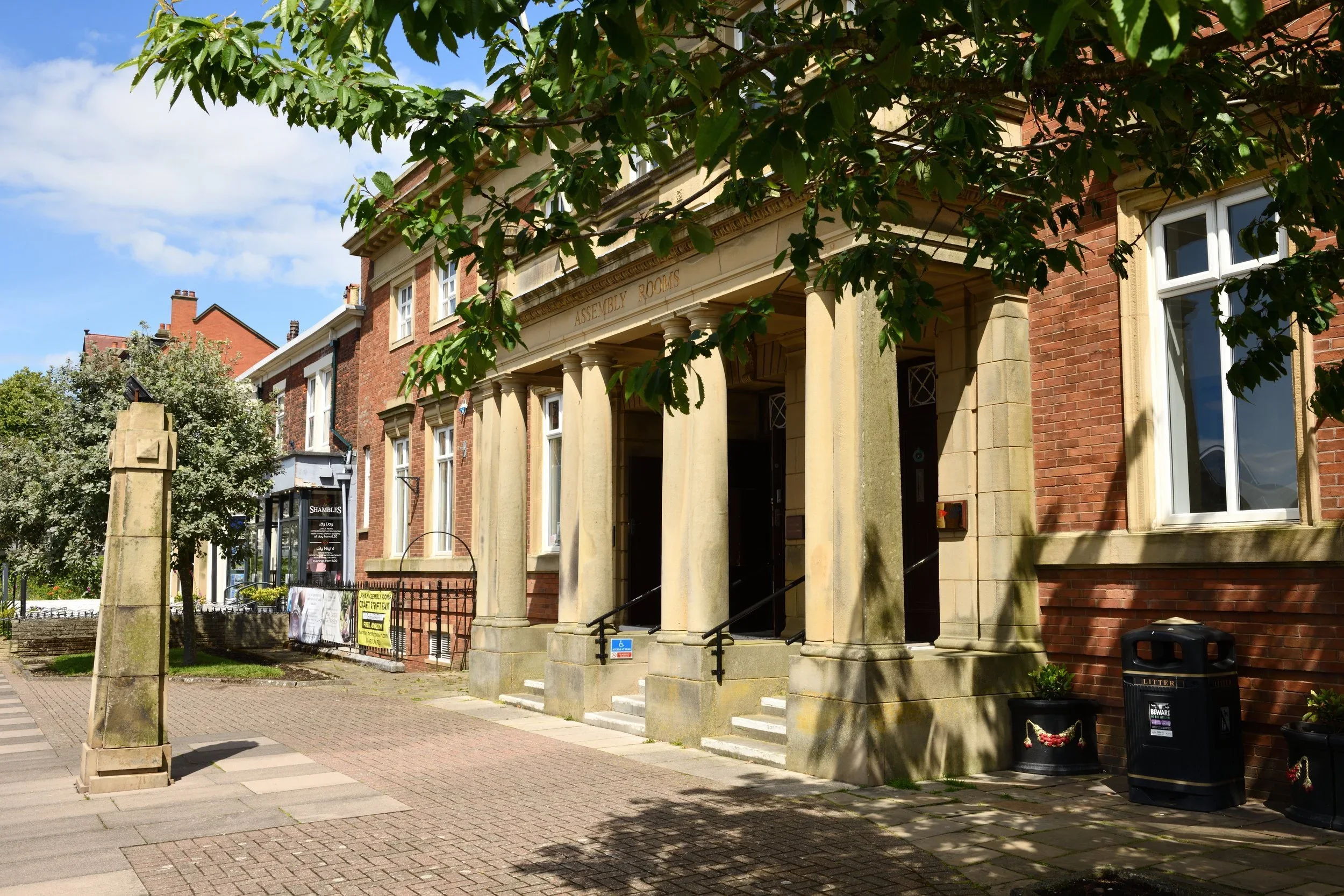 Street view of a historic brick building with columns and an inscription reading 'Assembly Rooms'. There are trees with green leaves partially obscuring the building, and a sidewalk with pedestrians and signage. The sky is partly cloudy.