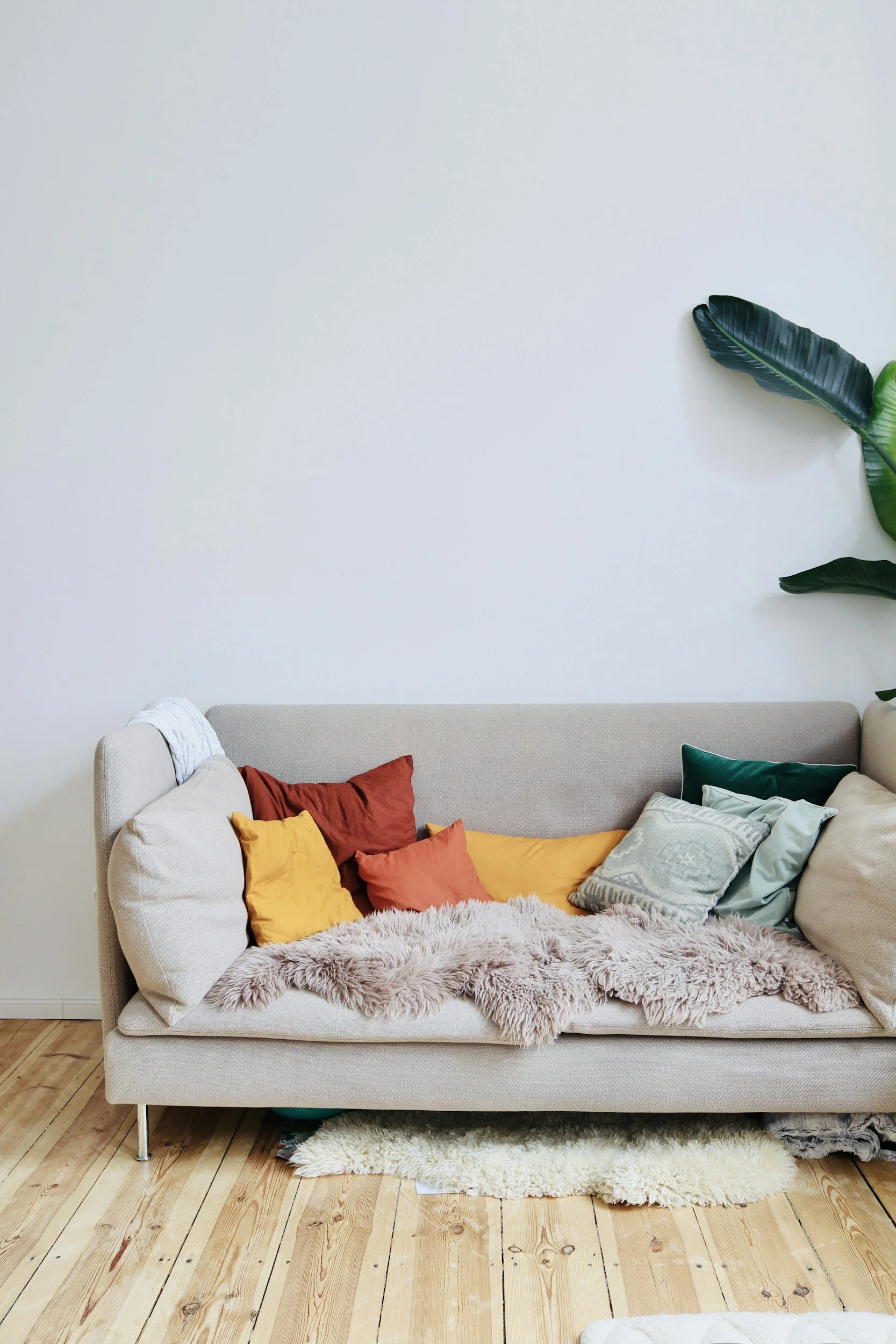 A beige sofa with colorful pillows, a fluffy beige blanket, situated in a room with light wooden flooring and a plain white wall with a large green leaf decoration.