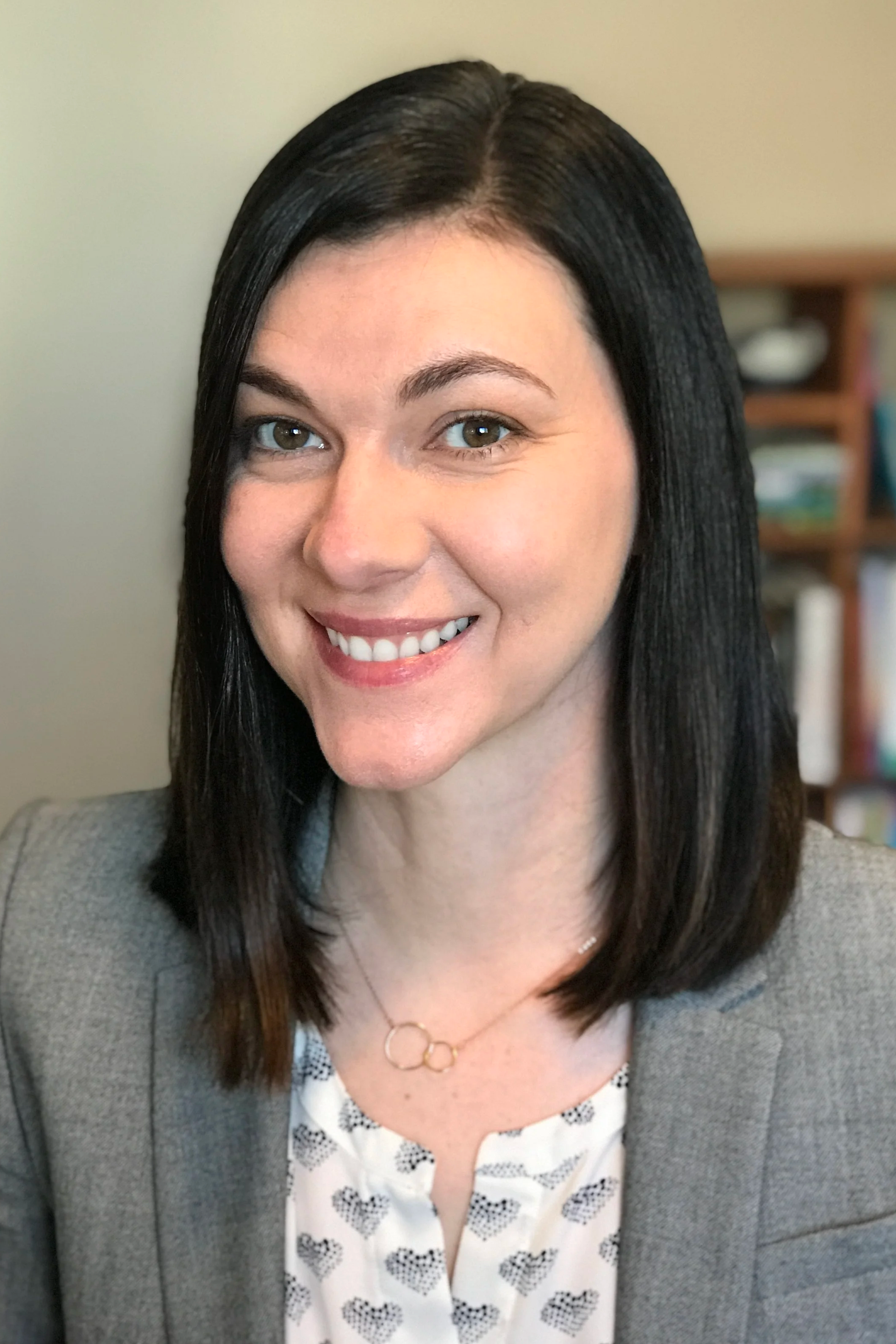 Headshot of a woman with dark brown, straight hair, smiling, wearing a gray blazer, white blouse with black hearts, and a gold necklace with three linked rings.