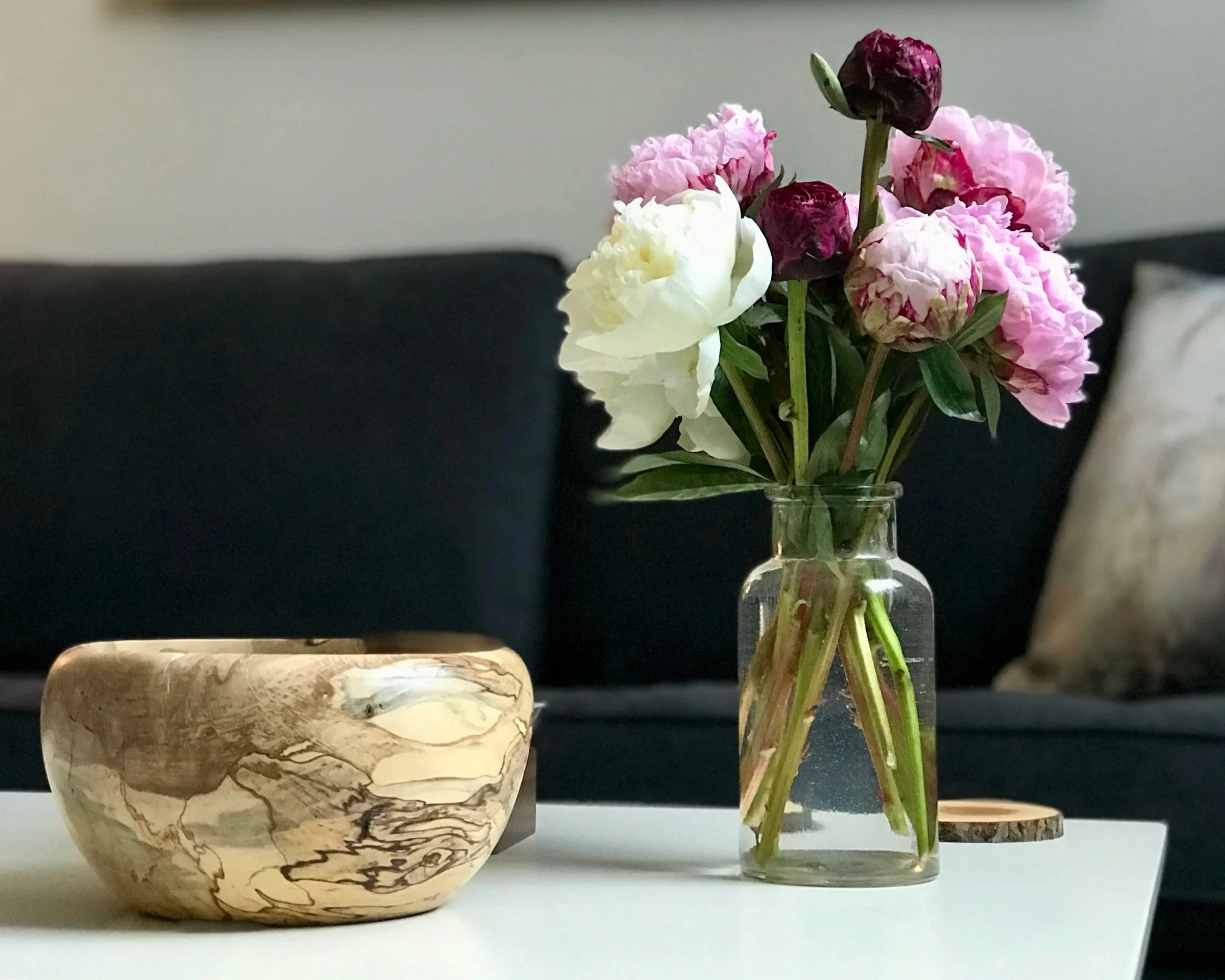 A clear glass vase holding pink, white, and dark purple flowers, placed next to a round, tan marble-patterned pot on a white surface with a dark blurry background.