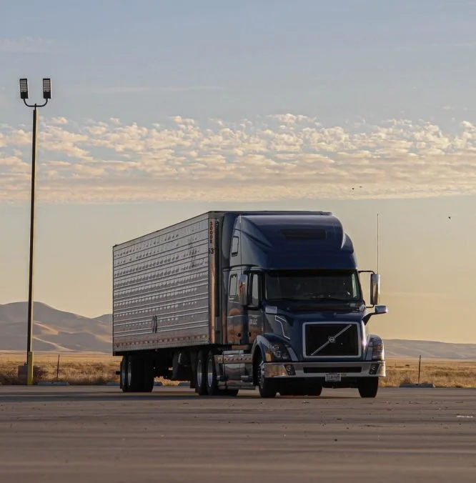 A large blue semi-truck parked in an open, flat area with mountains in the background and a tall streetlight nearby at sunset.