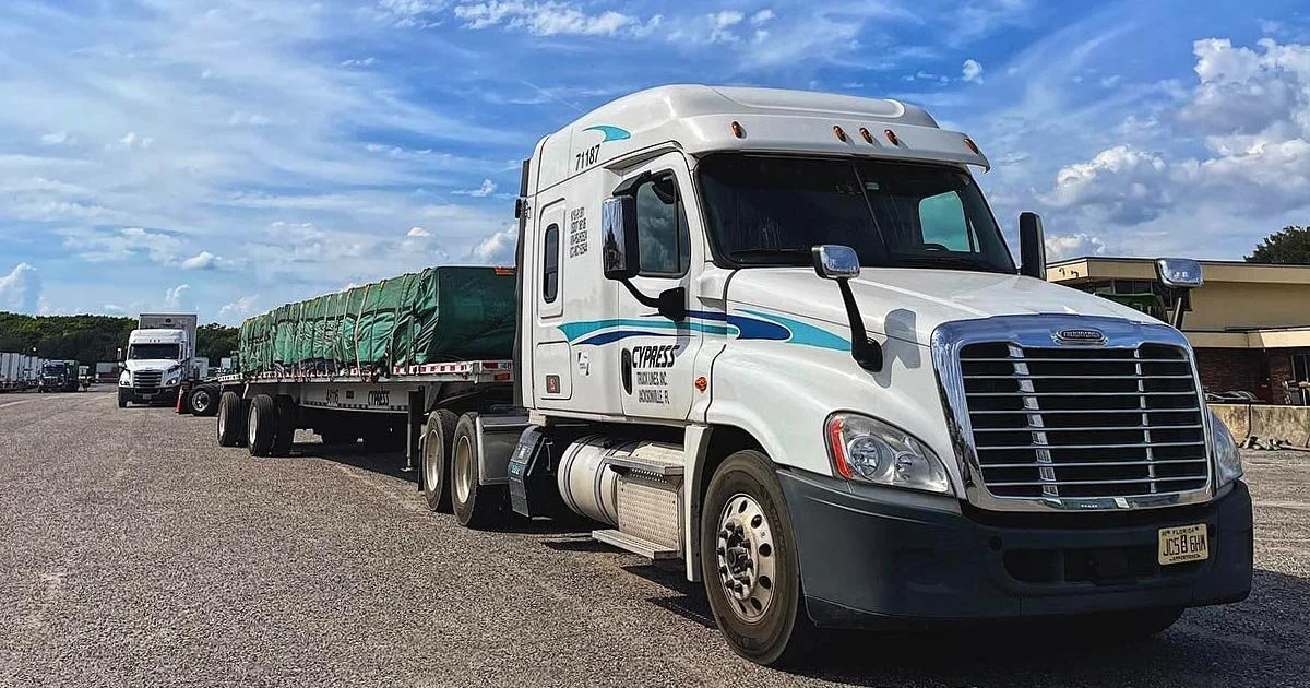 A white semi-truck with blue design and a flatbed trailer carrying green cargo, parked in a lot with a blue sky and scattered clouds.